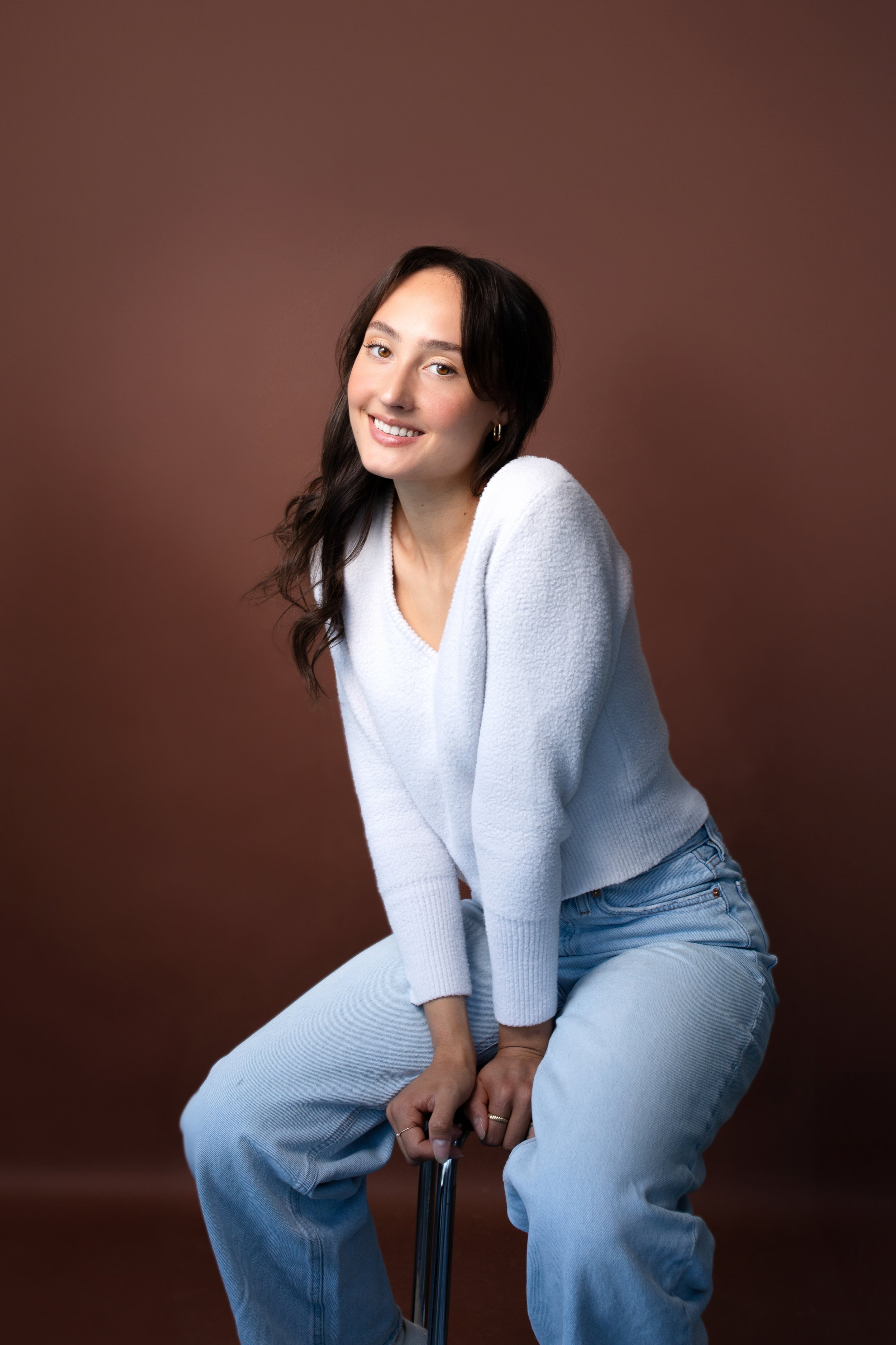 A young woman with dark, wavy hair sitting on a stool against a brown background. She is wearing a white sweater and light blue jeans, smiling with her hands clasped on her lap.