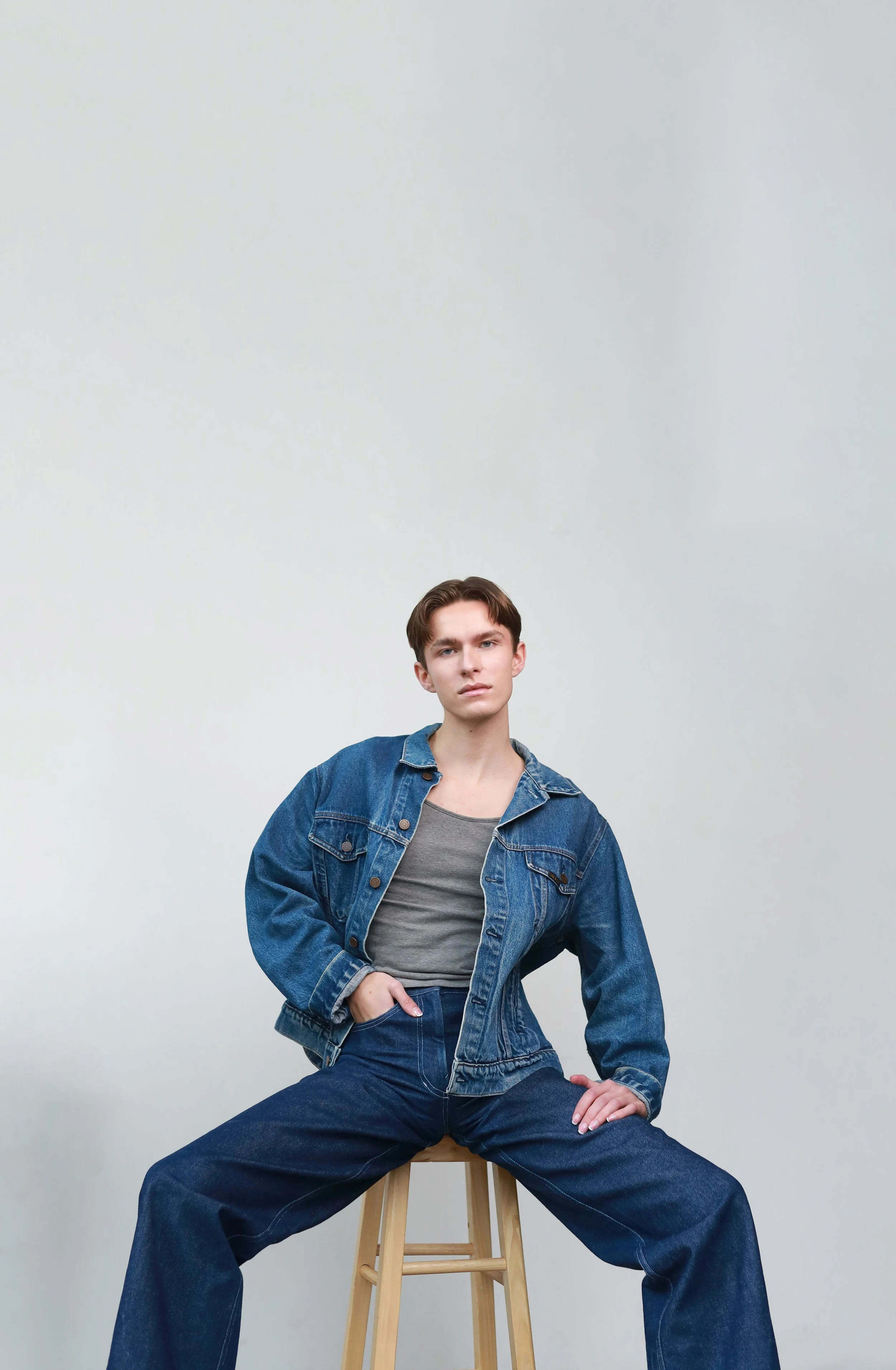 Young man in a denim jacket and jeans sitting on a stool with legs wide apart in front of a plain white wall.