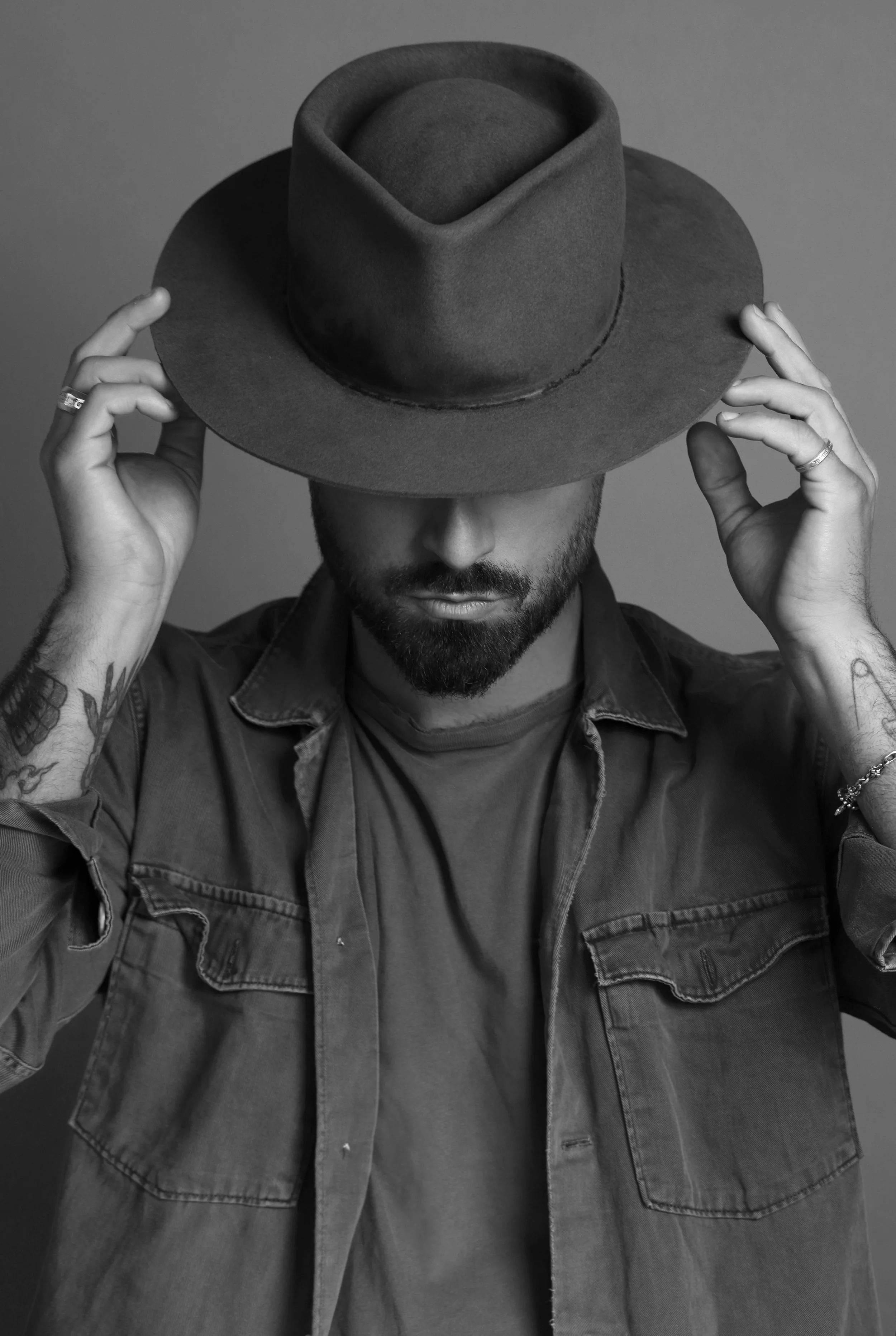 A man with tattoos on his arms wearing a denim jacket, holding a wide-brimmed hat over his face, in a black and white photograph.