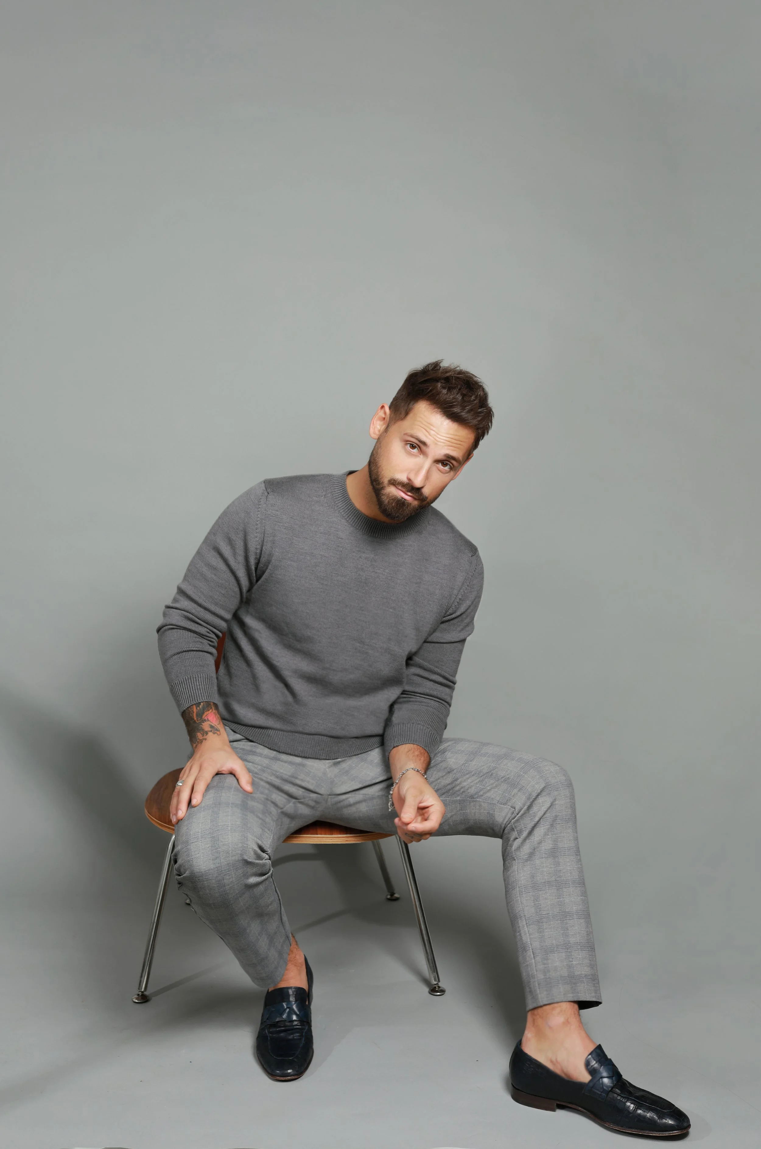 A man with a beard and mustache, wearing a gray sweater and gray checkered pants, sits on a wooden chair against a plain gray background. He has short brown hair, tattoos on his left arm, and is wearing black loafers. He is leaning slightly forward w