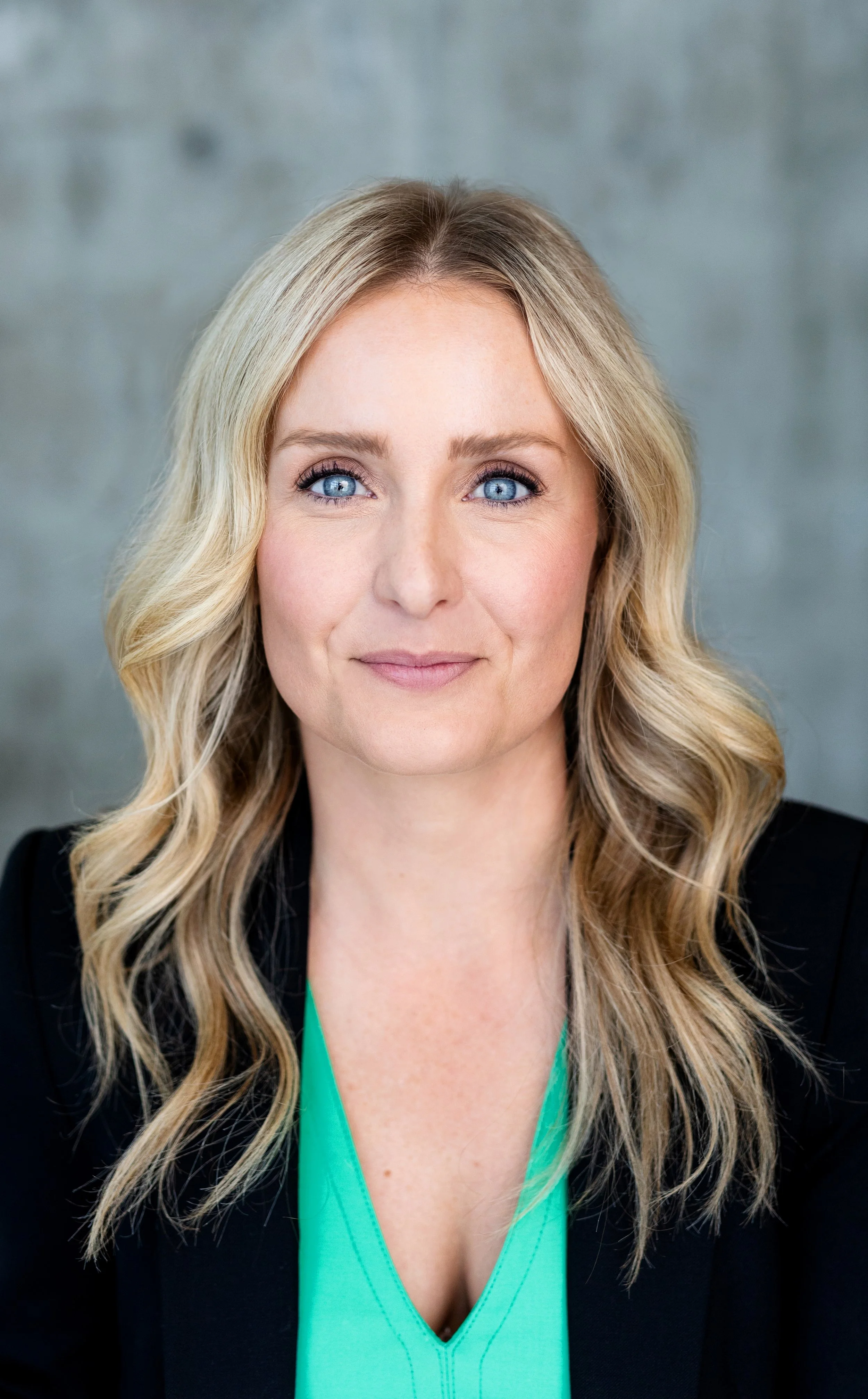 A woman with long, wavy blonde hair and blue eyes is smiling gently, wearing a black blazer over a green top, against a blurred gray background.