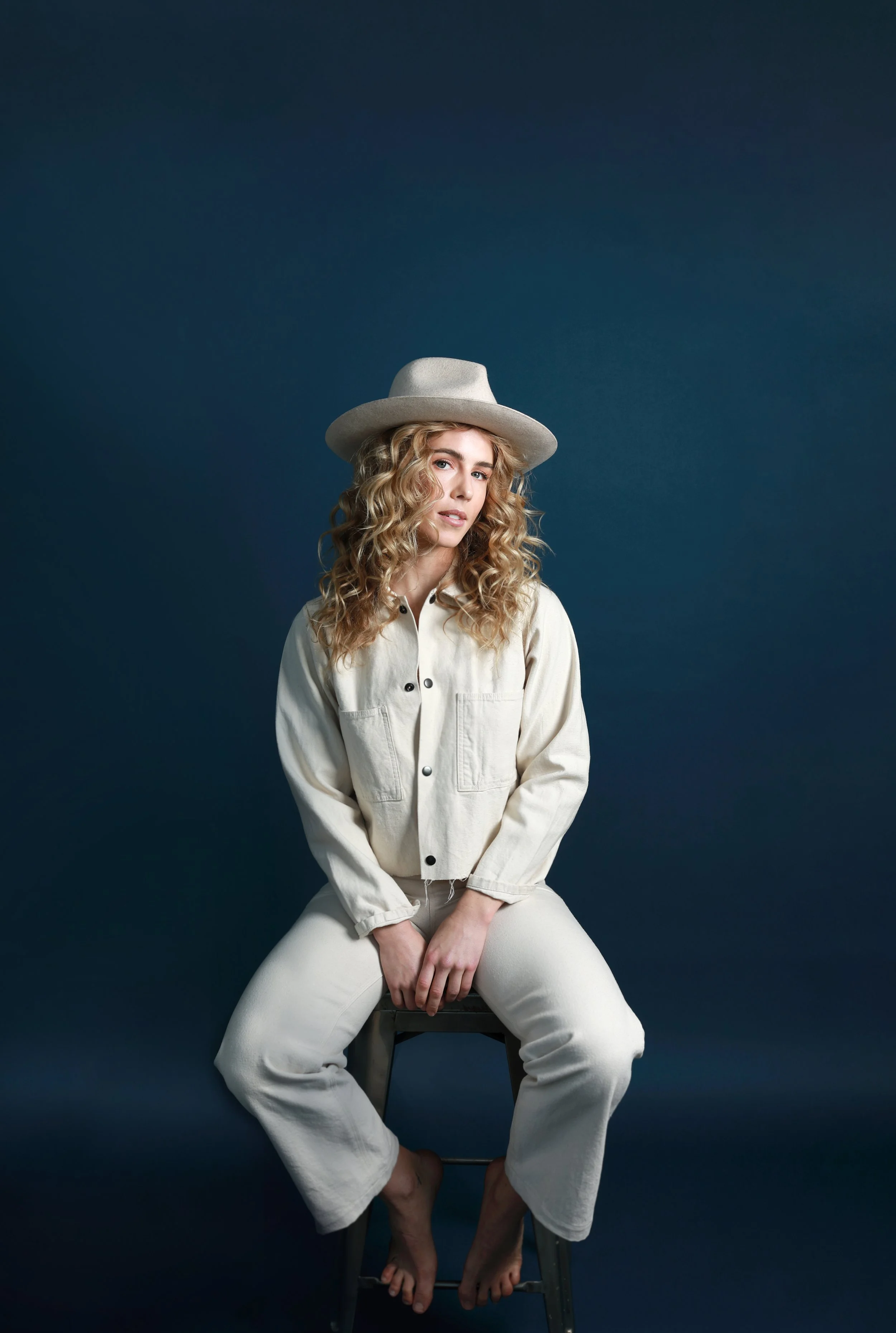 A woman with blonde curly hair wearing a beige hat and beige button-up outfit, sitting on a wooden stool against a dark blue background.