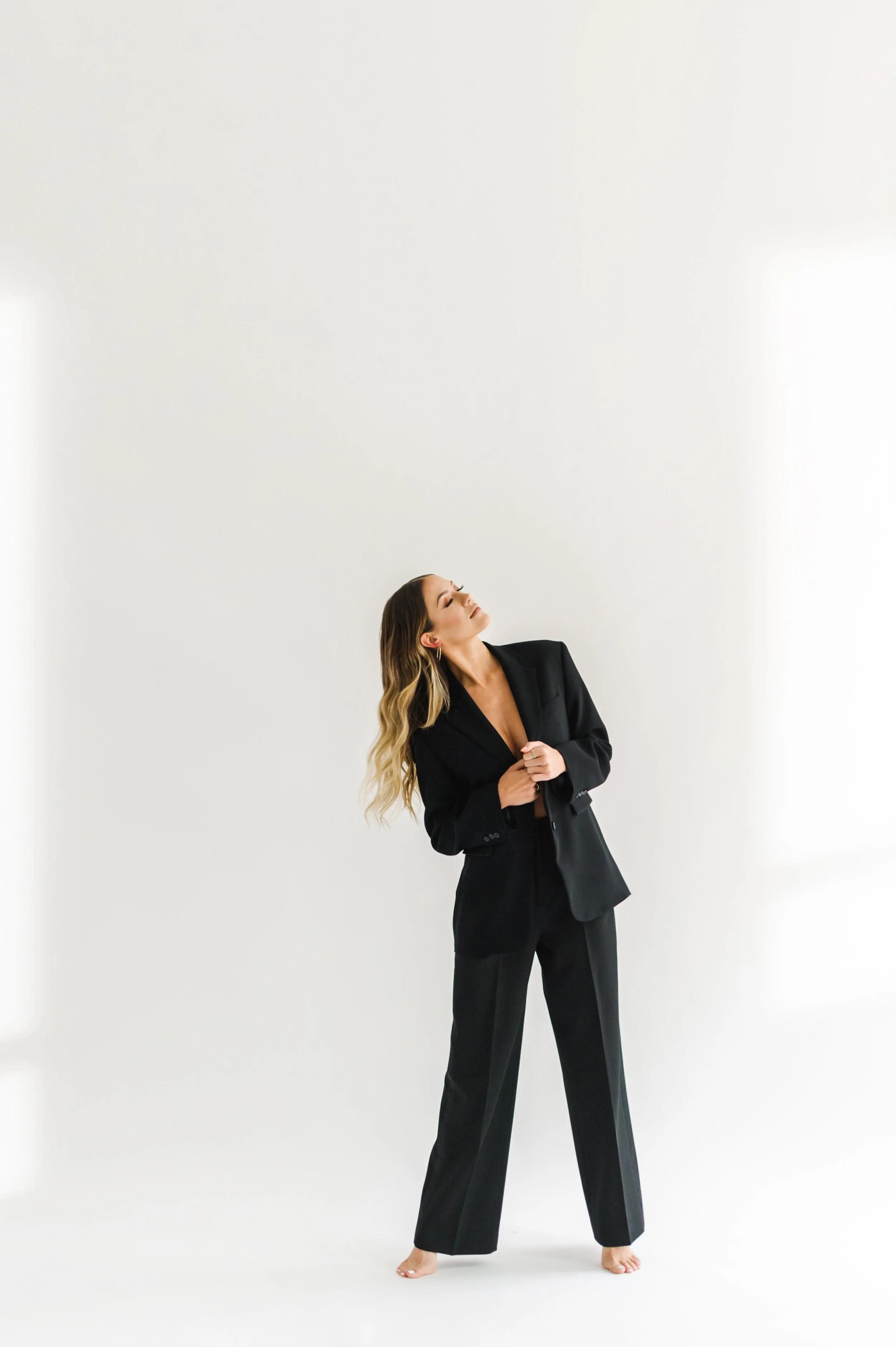 A woman with long wavy hair wearing a black suit, standing with one foot slightly forward and her head tilted back on a plain white background.