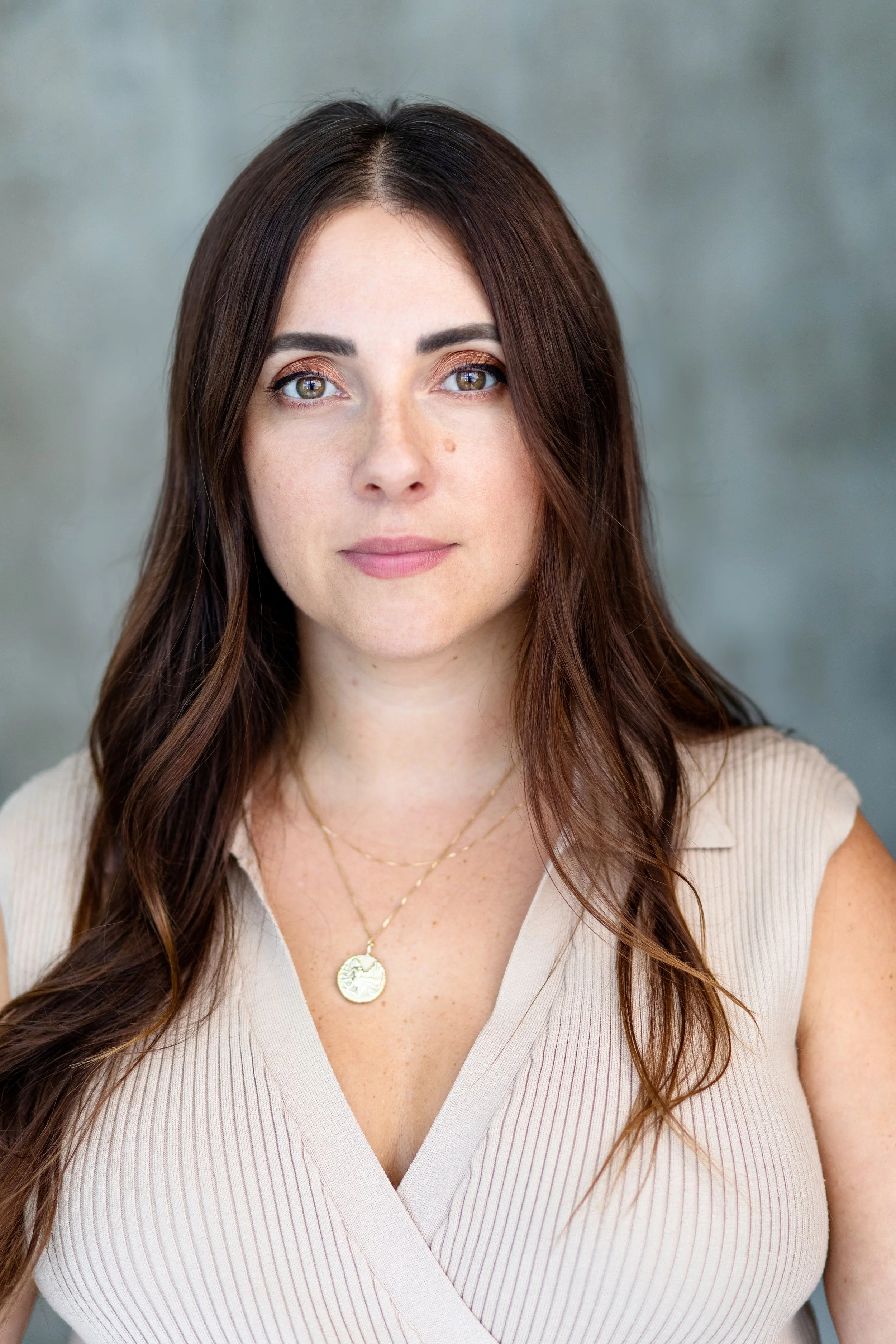 Headshot of a woman with long brown hair and light green eyes, wearing makeup and a beige V-neck top with a gold necklace, standing against a gray background.