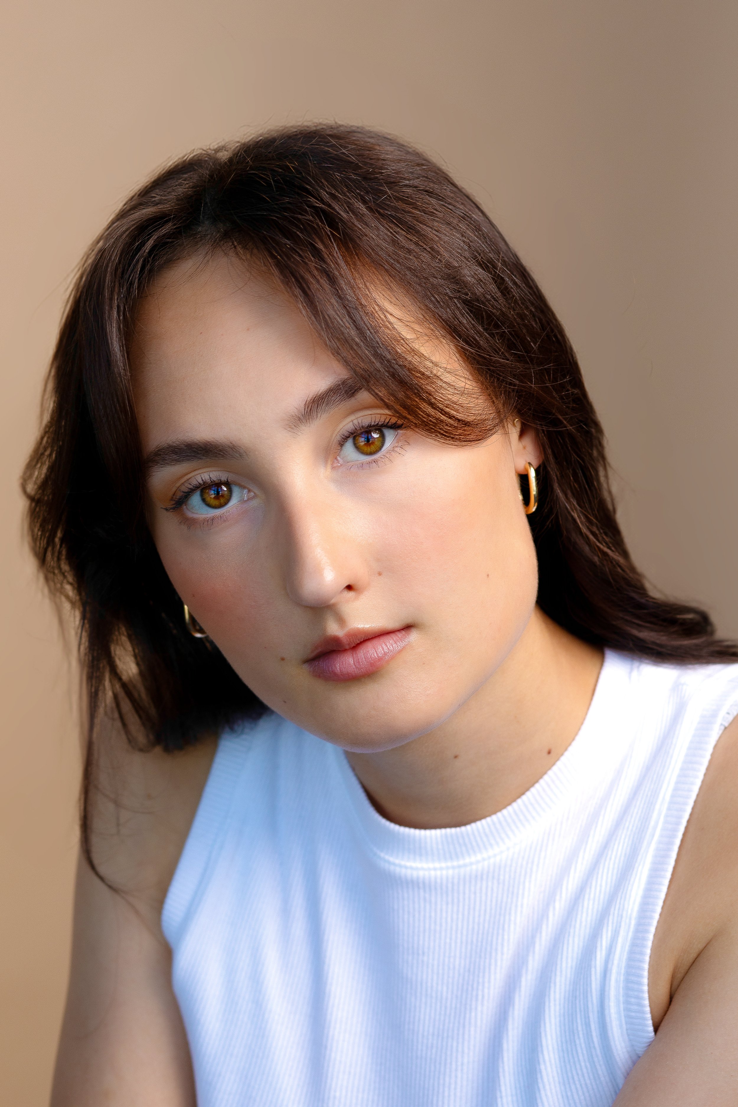 Close-up of a young woman with brown hair, hazel eyes, and light makeup, wearing a white sleeveless top and gold hoop earrings, posing against a neutral beige background.