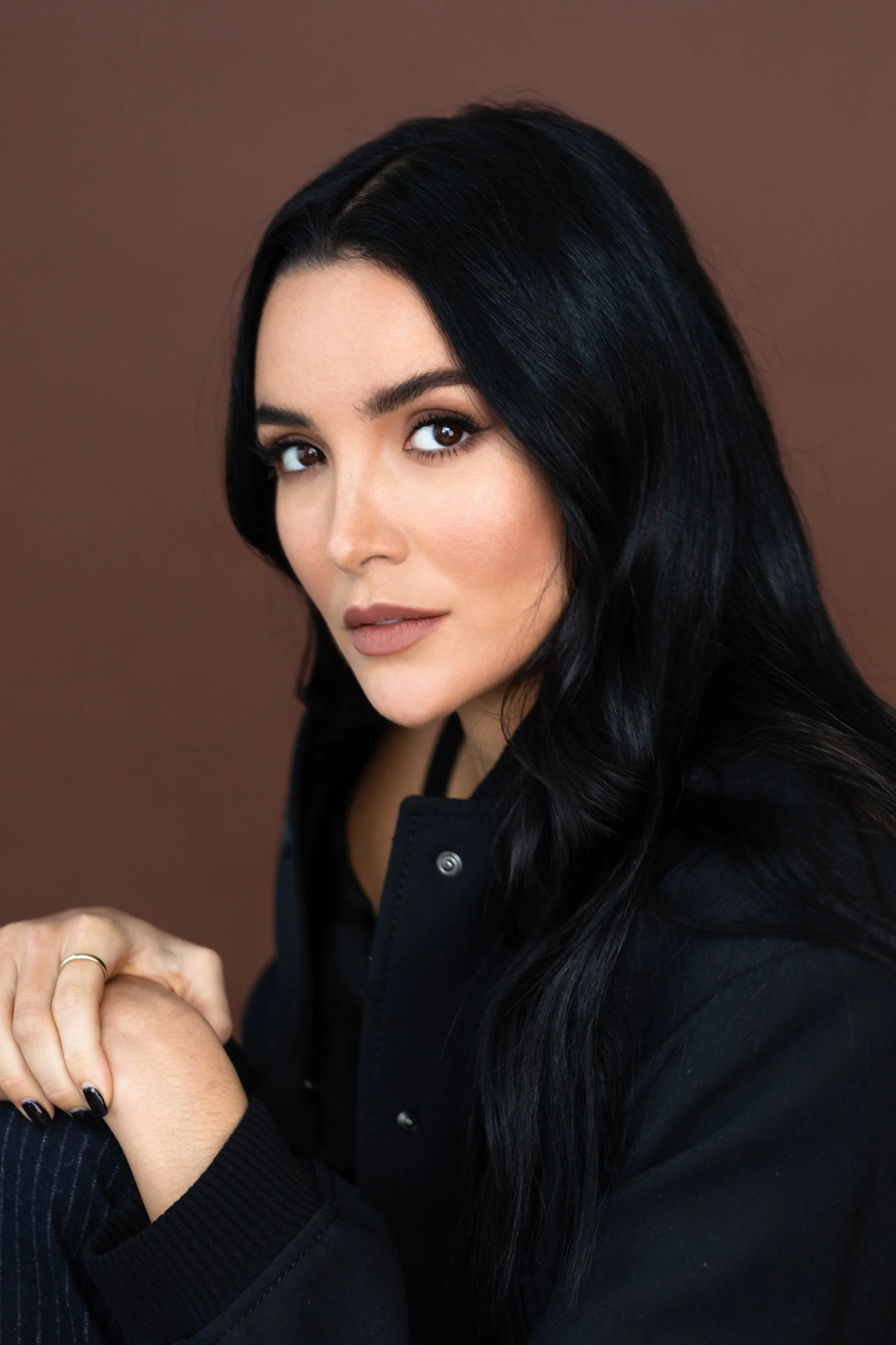 Close-up portrait of a woman with long black hair, brown eyes, and light makeup, wearing a black jacket with silver buttons, against a brown background.