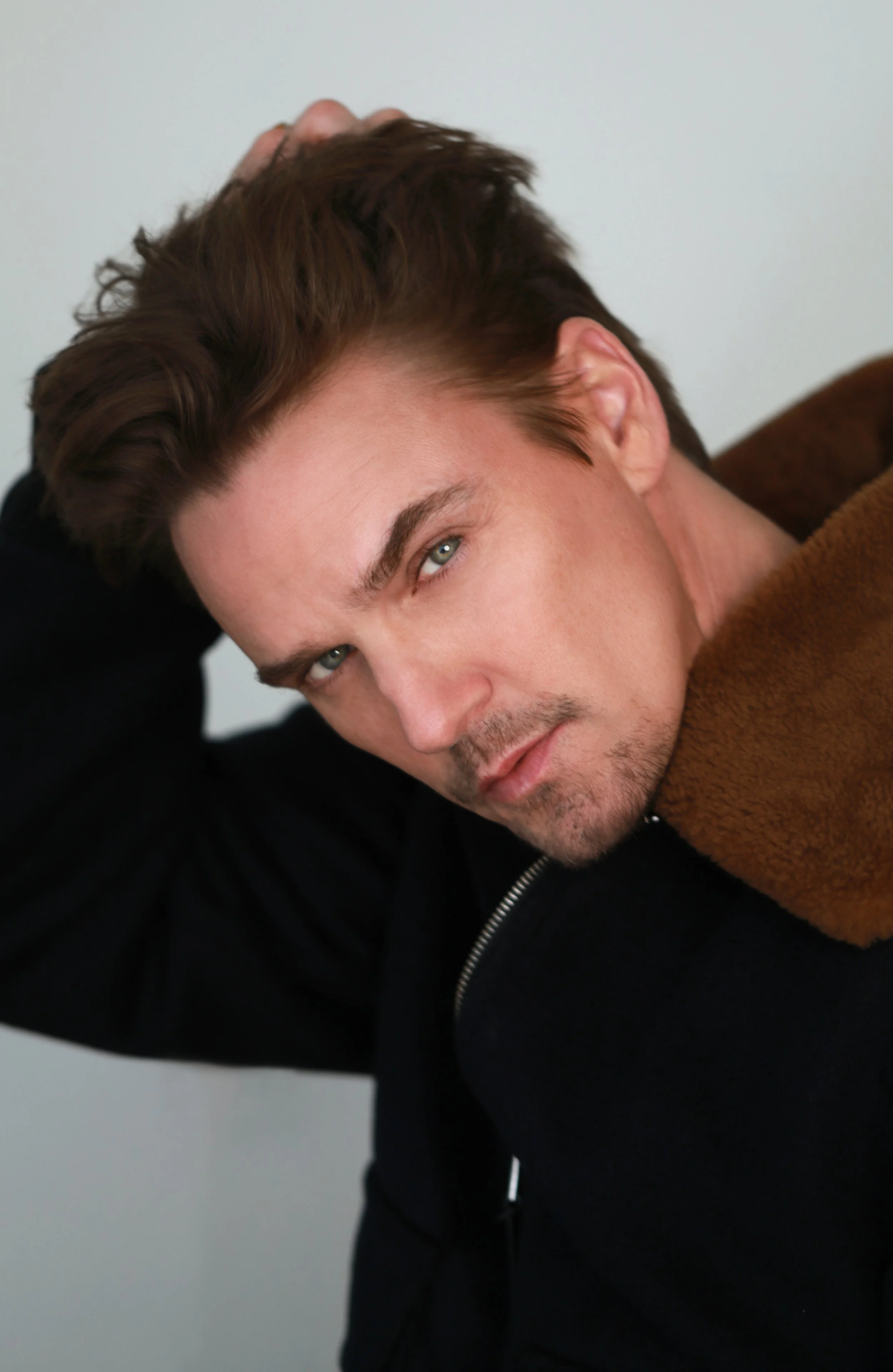 A young man with short, wavy brown hair and green eyes lying on his side, looking into the camera, wearing a brown coat and black hoodie, against a simple light-colored background.