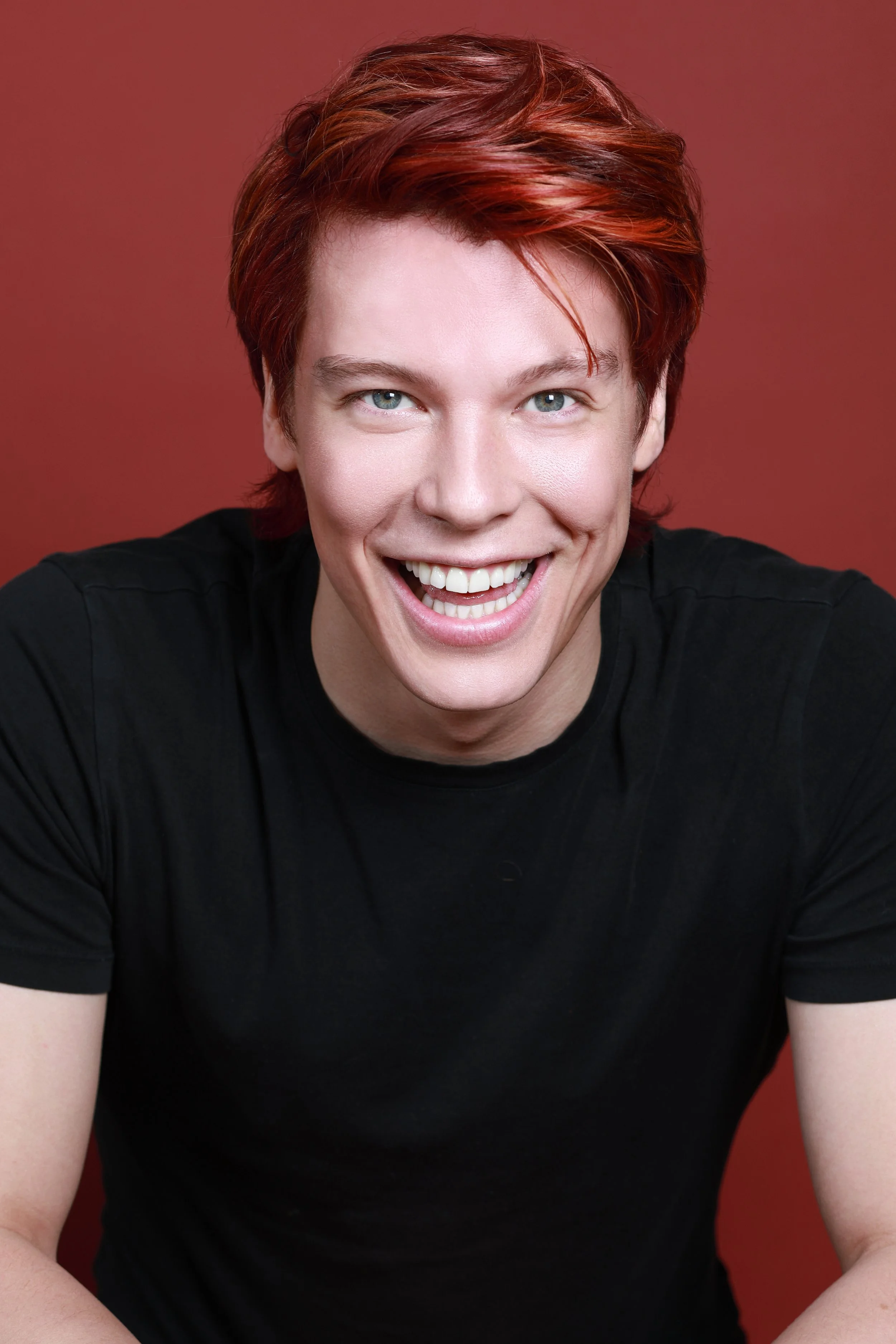 A person with short red hair and blue eyes, smiling widely, wearing a black shirt against a reddish-brown background.