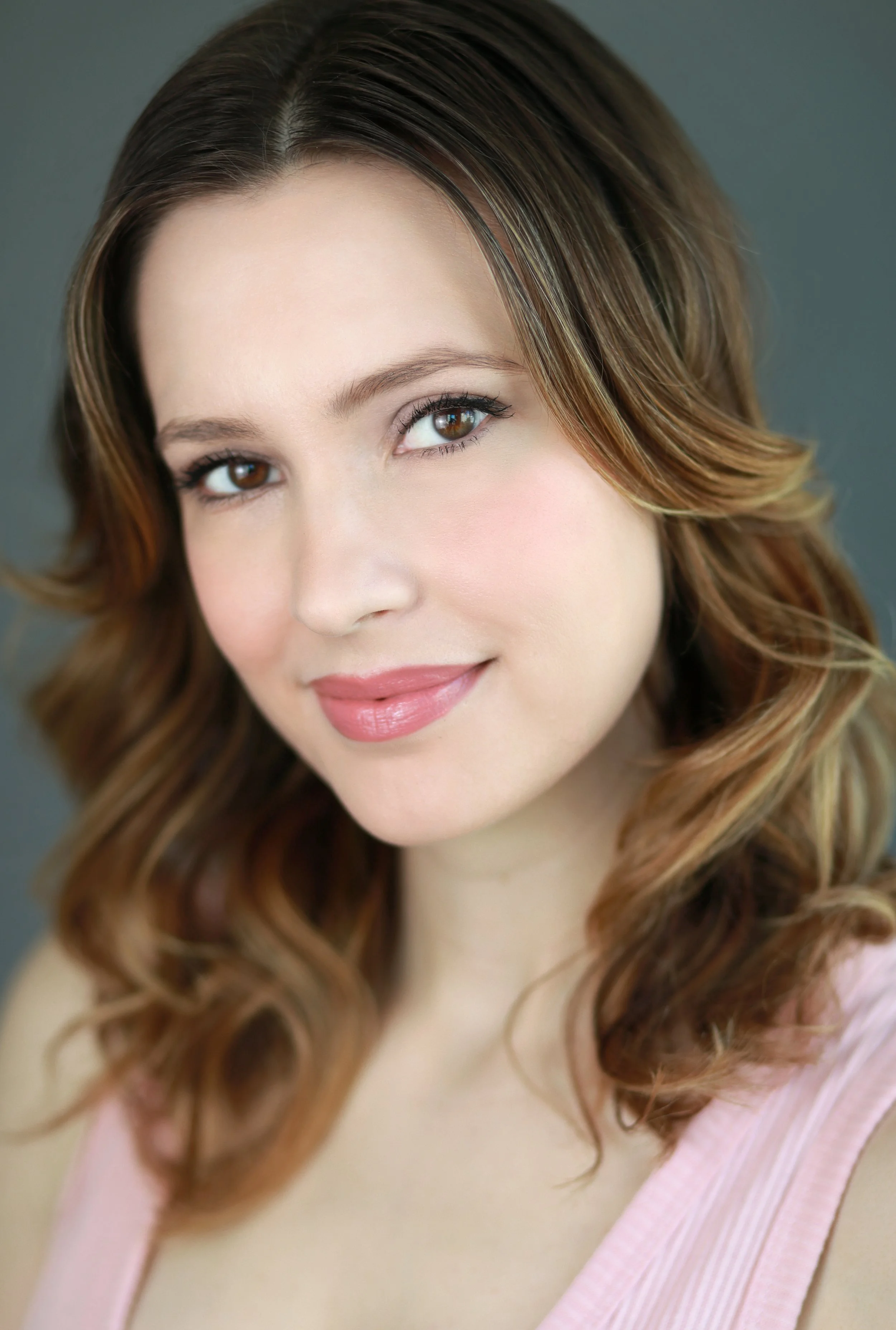 Close-up of a young woman with brown hair styled in loose waves, wearing a pink top with a textured strap.