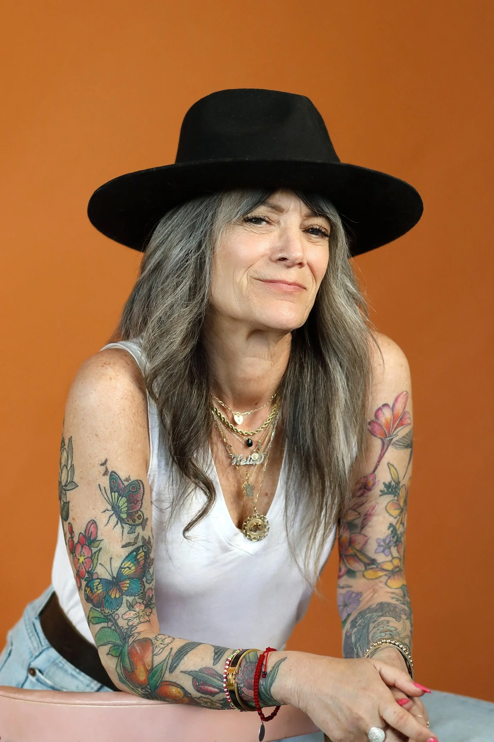 A smiling woman with long gray hair, tattoos on her arms, wearing a black wide-brimmed hat, layered necklaces, and a white tank top, leaning on a pink table with an orange background.