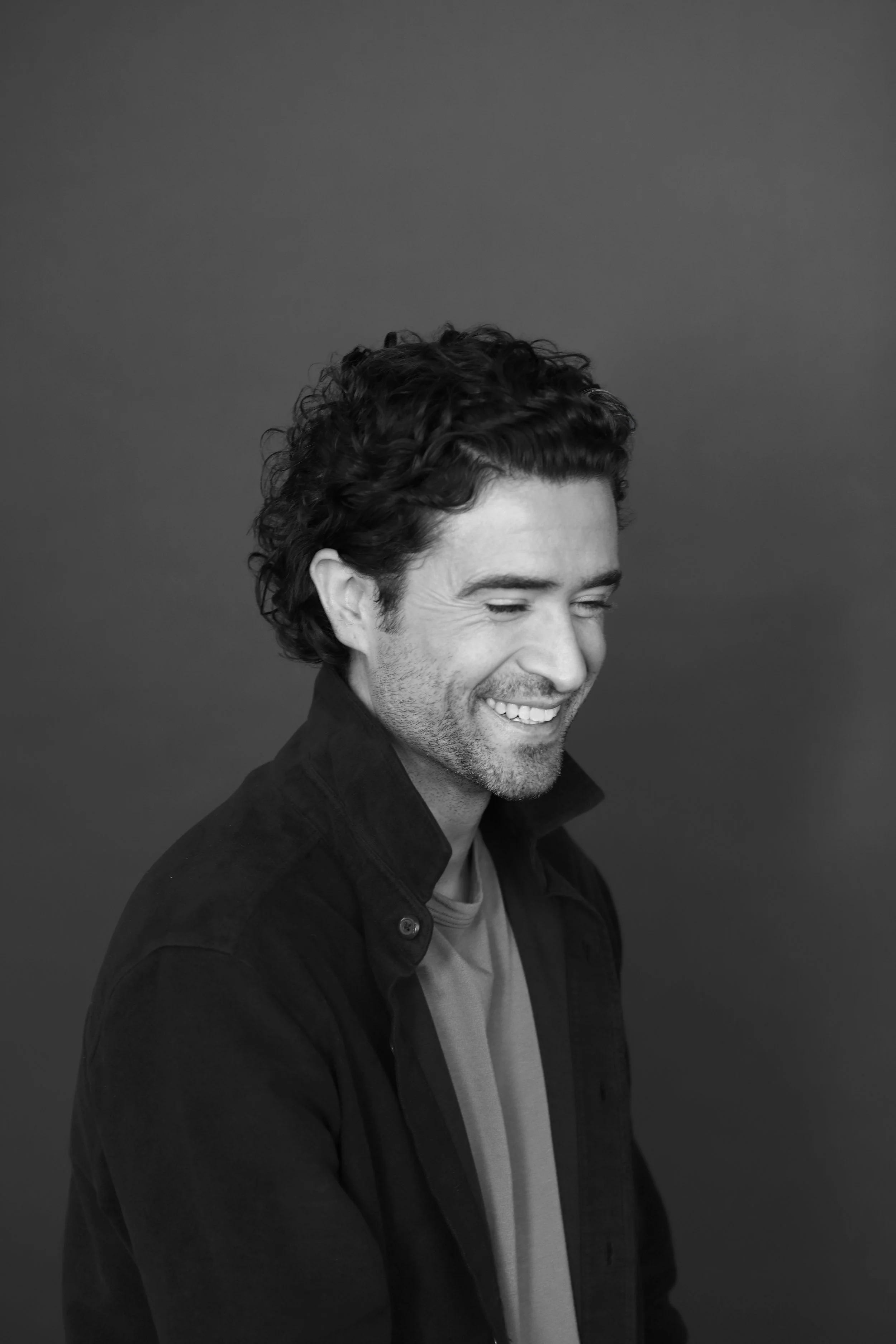 A smiling man with curly dark hair, wearing a black jacket and a t-shirt, shot in black and white against a plain background.