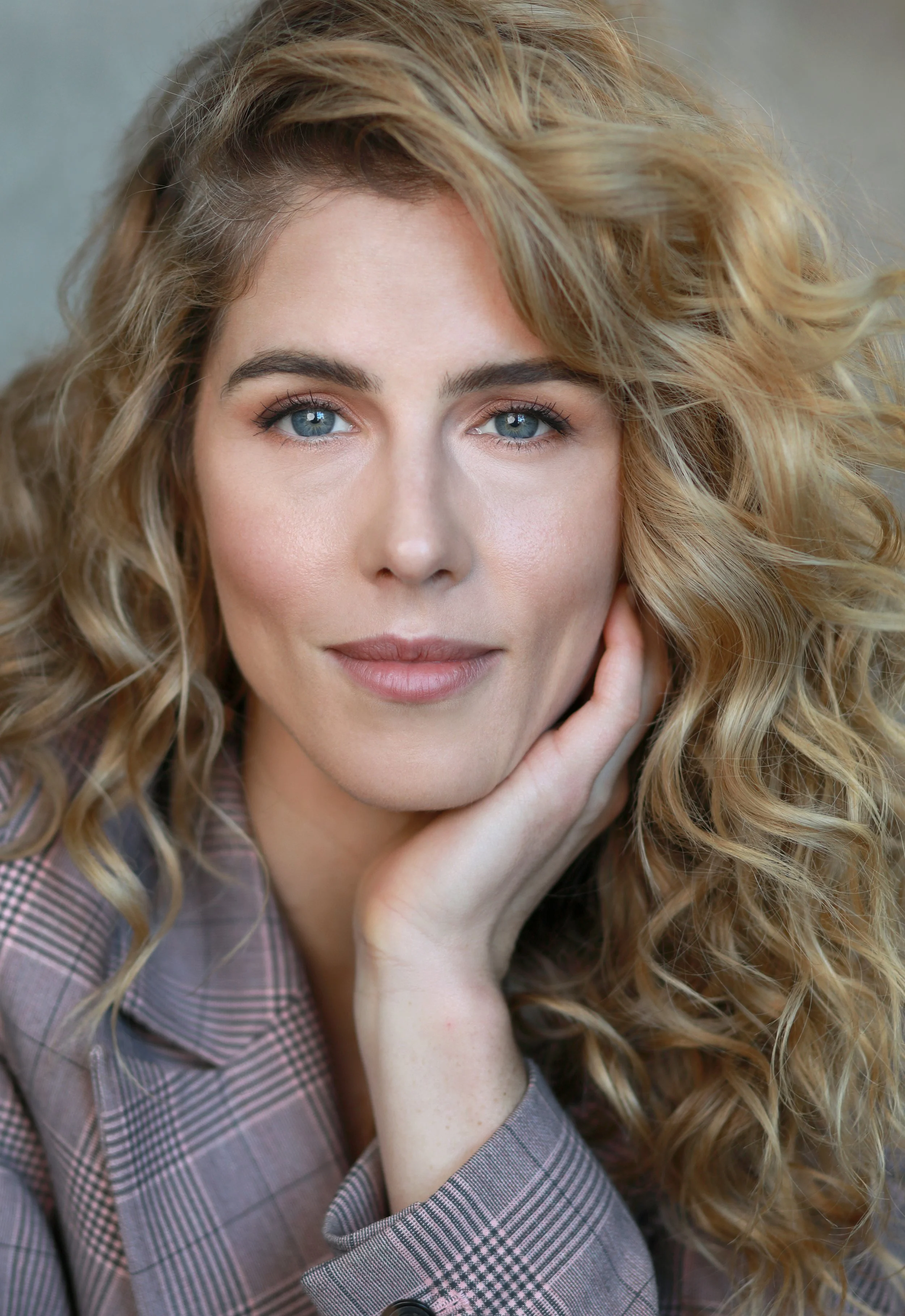 Close-up portrait of a woman with curly blonde hair and blue eyes, resting her chin on her hand, wearing a pink plaid blazer.
