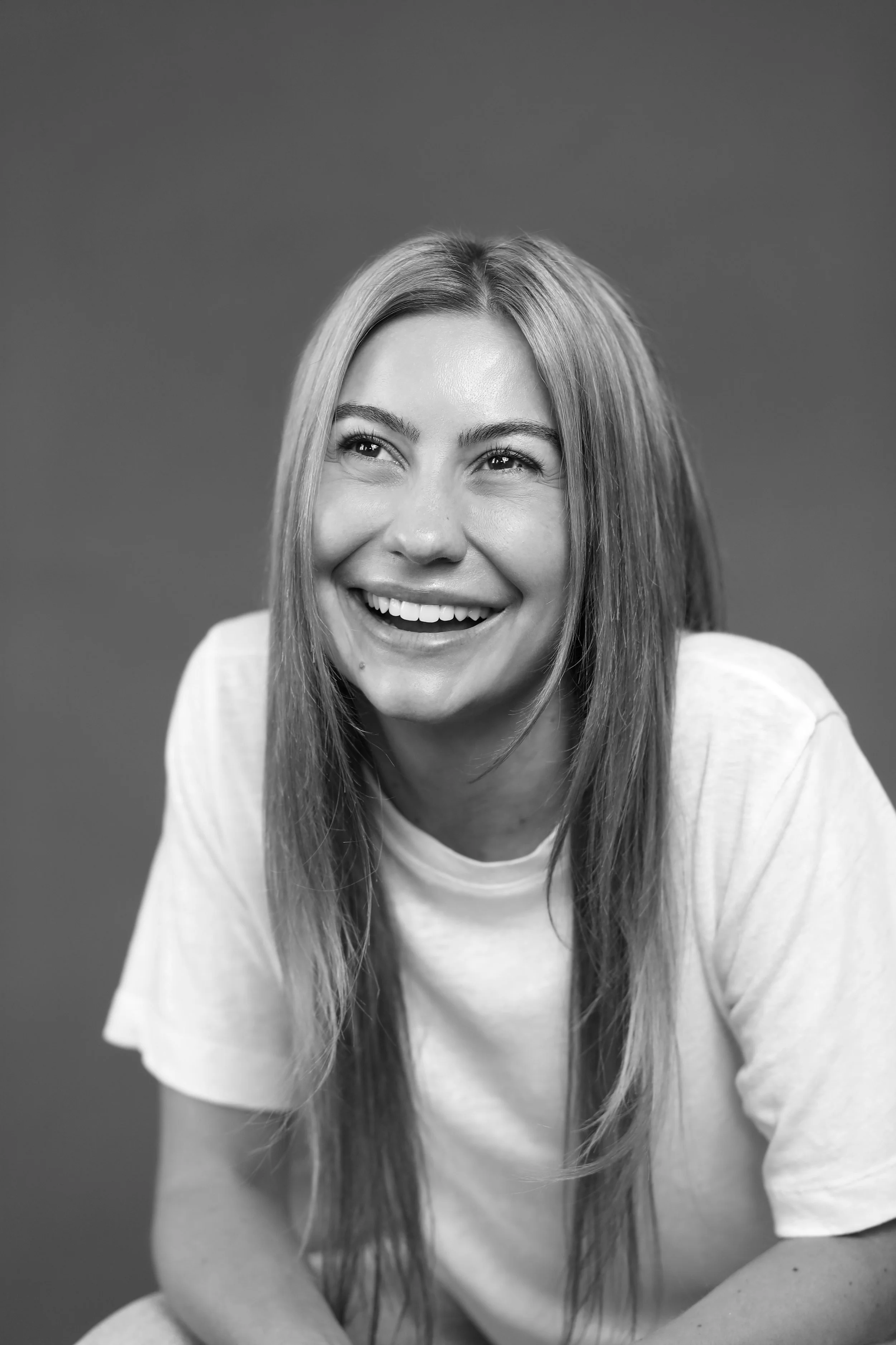 Black and white photo of a young woman smiling and looking upward, wearing a plain light-colored t-shirt, with long hair and a blurred background.