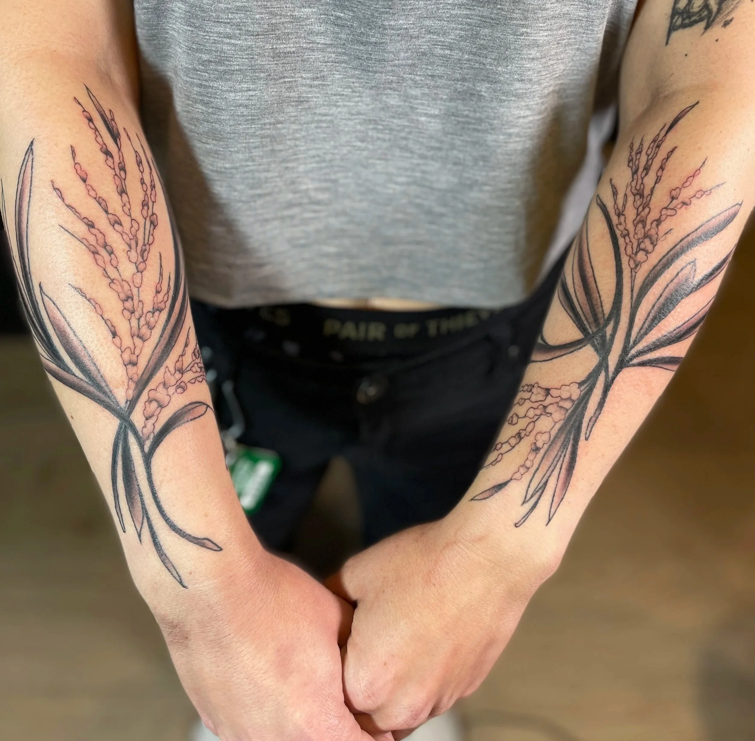 Person with new botanical tattoos of leaves and sprigs on their arms, wearing a gray shirt and black pants.