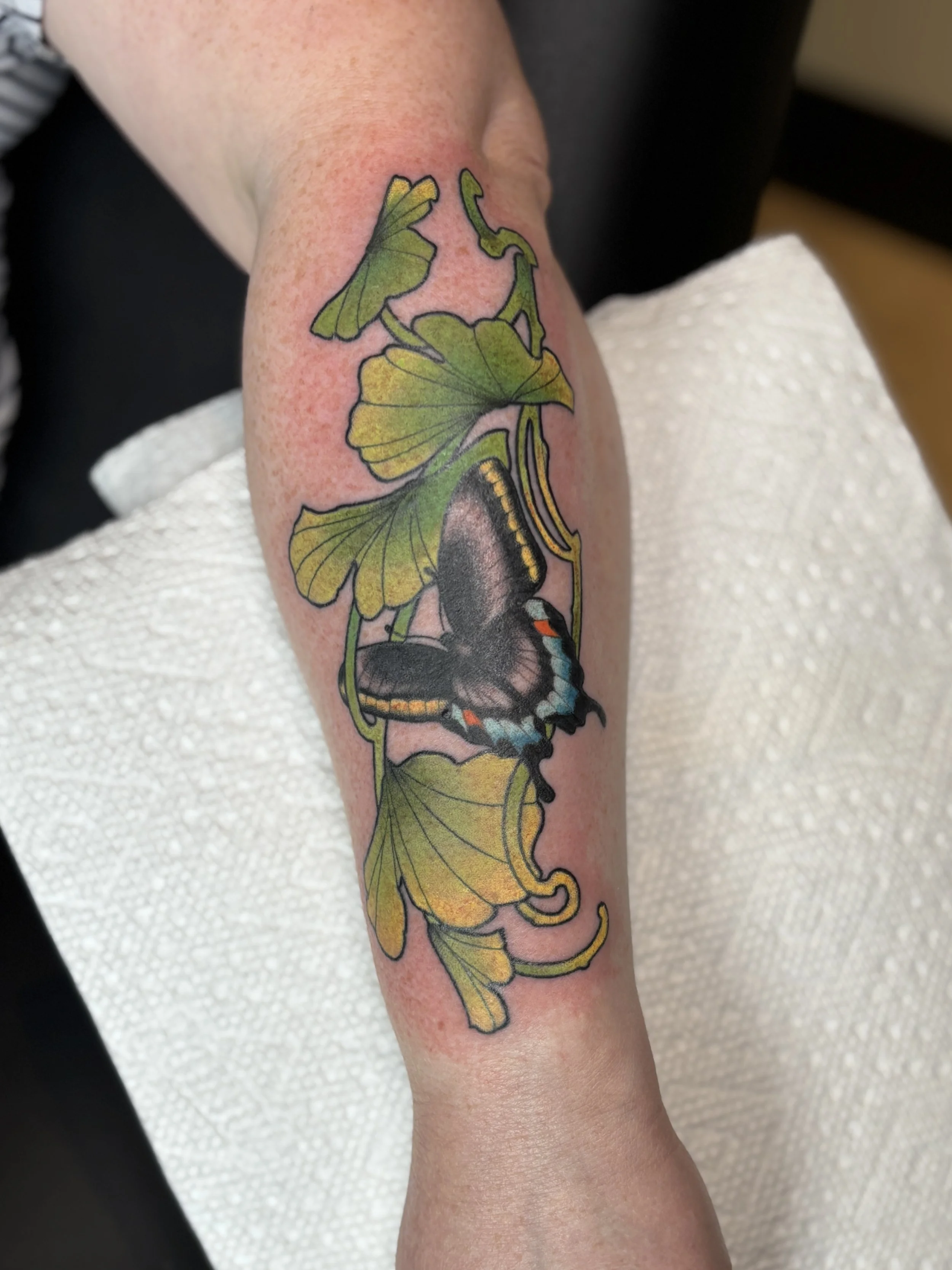 Colorful tattoo of a butterfly and leaves on a person's forearm.