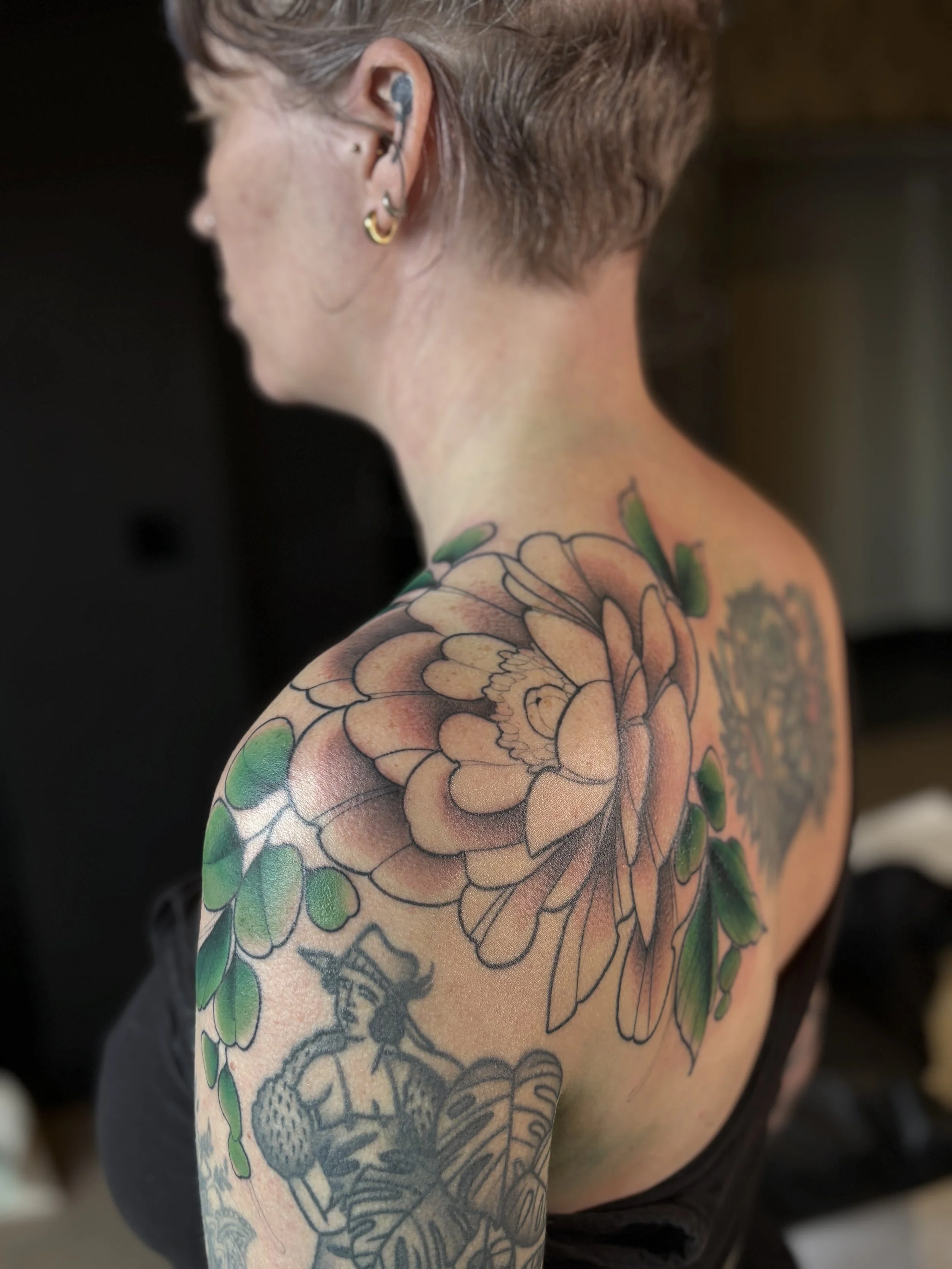 A woman with short hair and multiple earrings showcases colorful floral tattoos on her shoulder and upper arm, including a large peach-colored flower with green leaves.