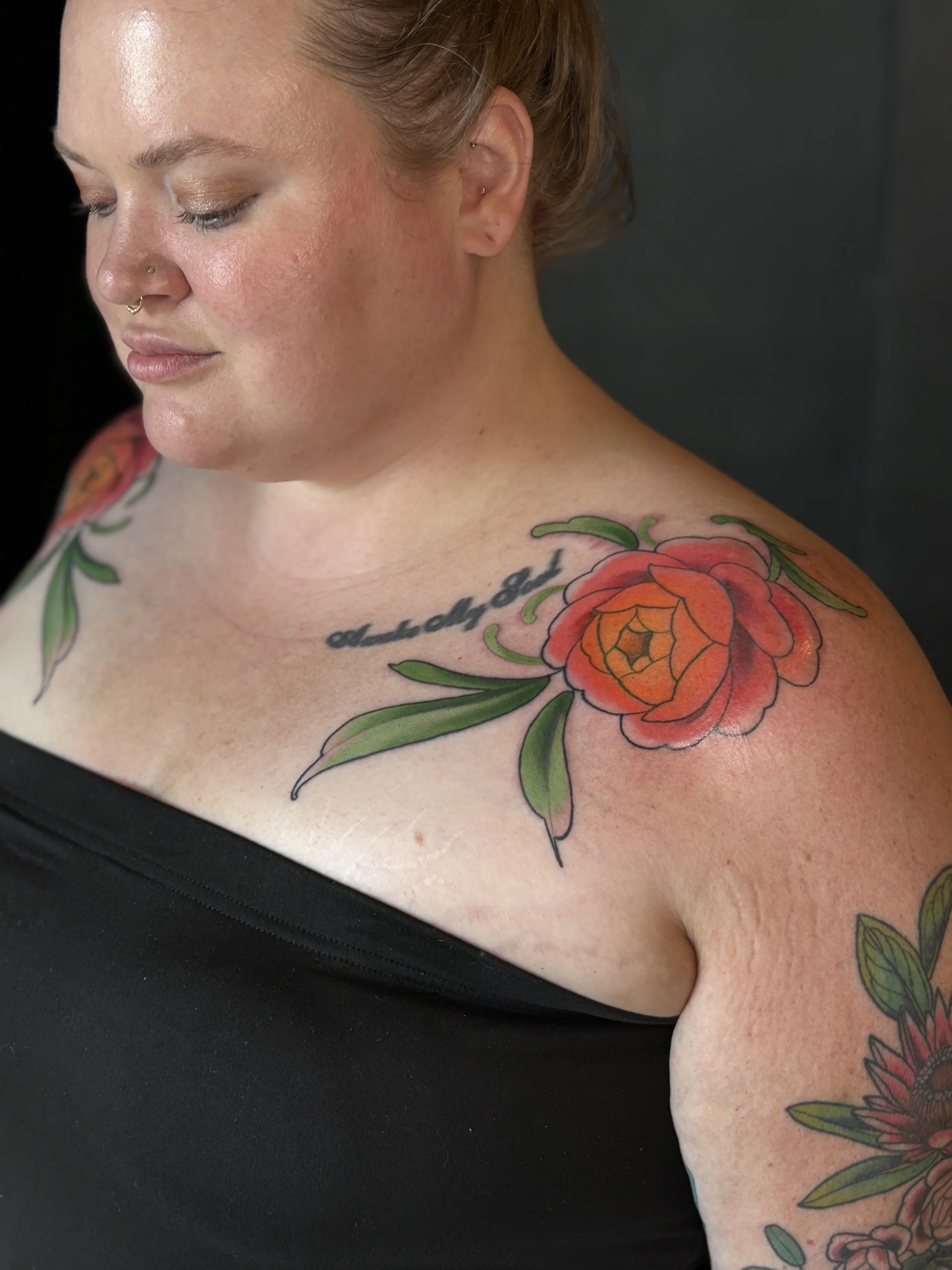 Close-up of a woman with tattoos on her shoulder and chest, including a large red and orange flower with green leaves, and a part of a tattooed quote.