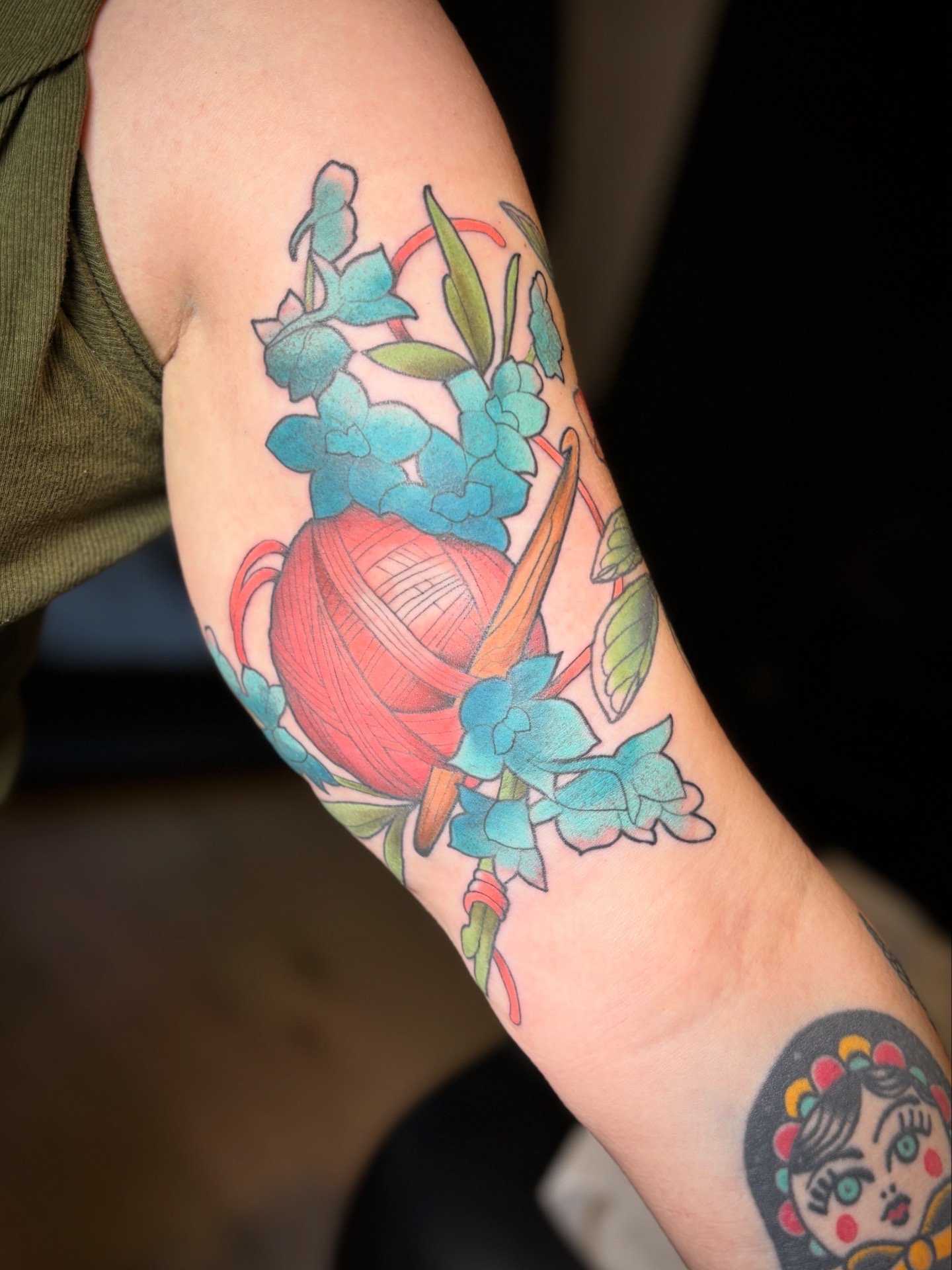 Colorful tattoo of a ball of yarn wrapped with knitting needles, surrounded by blue flowers and green leaves on a person's arm.
