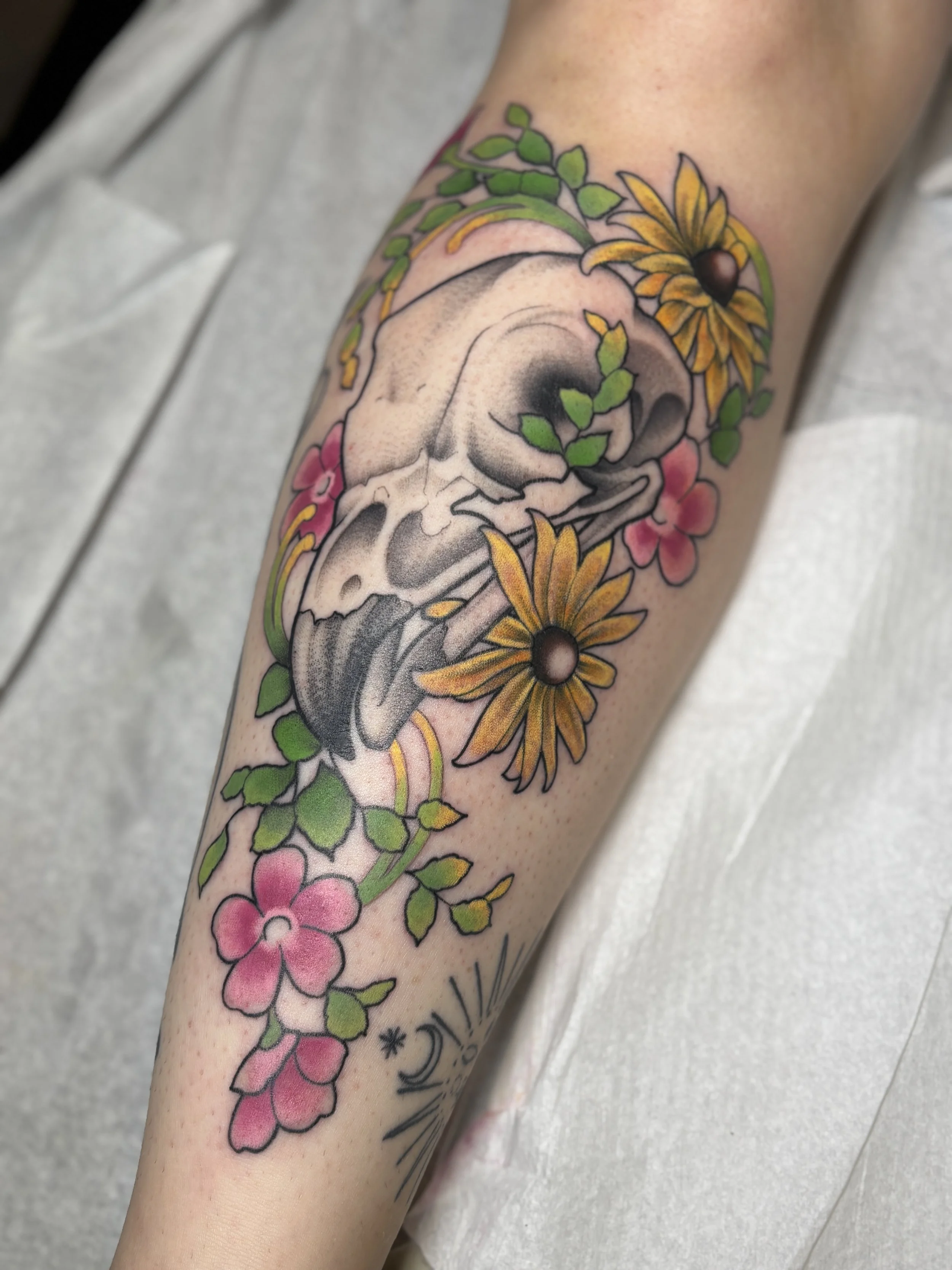Tattoo of a skull surrounded by yellow daisies, pink flowers, green leaves, and decorative elements on a person's arm.