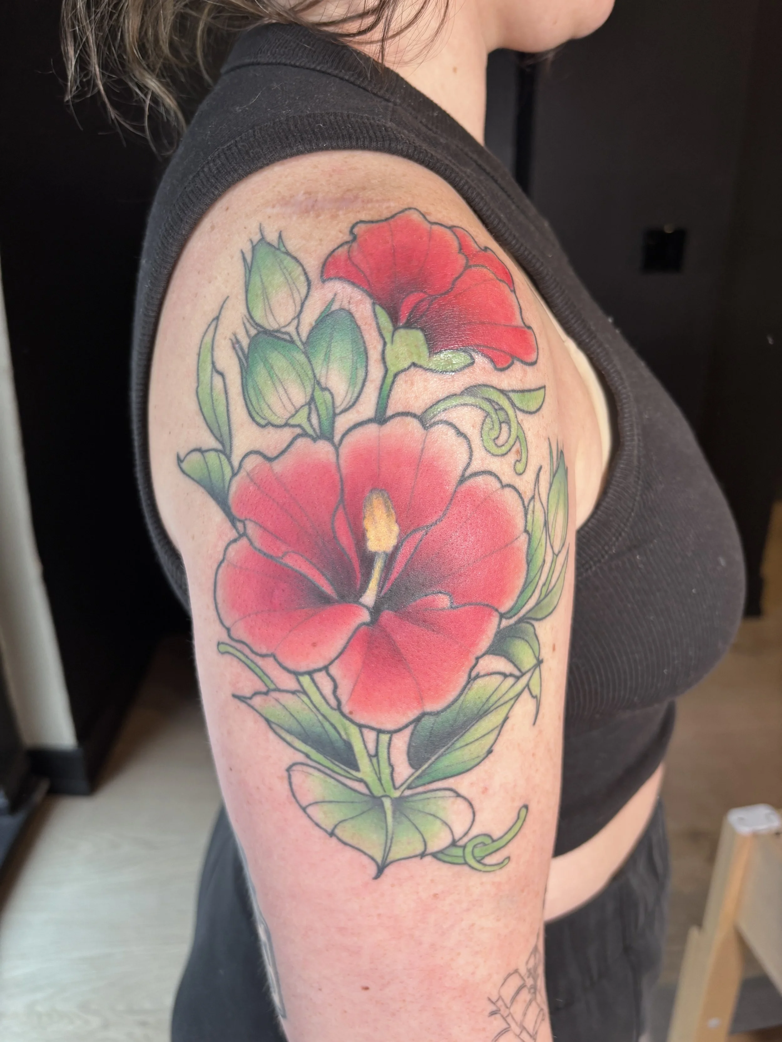 Colorful tattoo of a red hibiscus flower with green leaves and buds on a woman's upper arm.