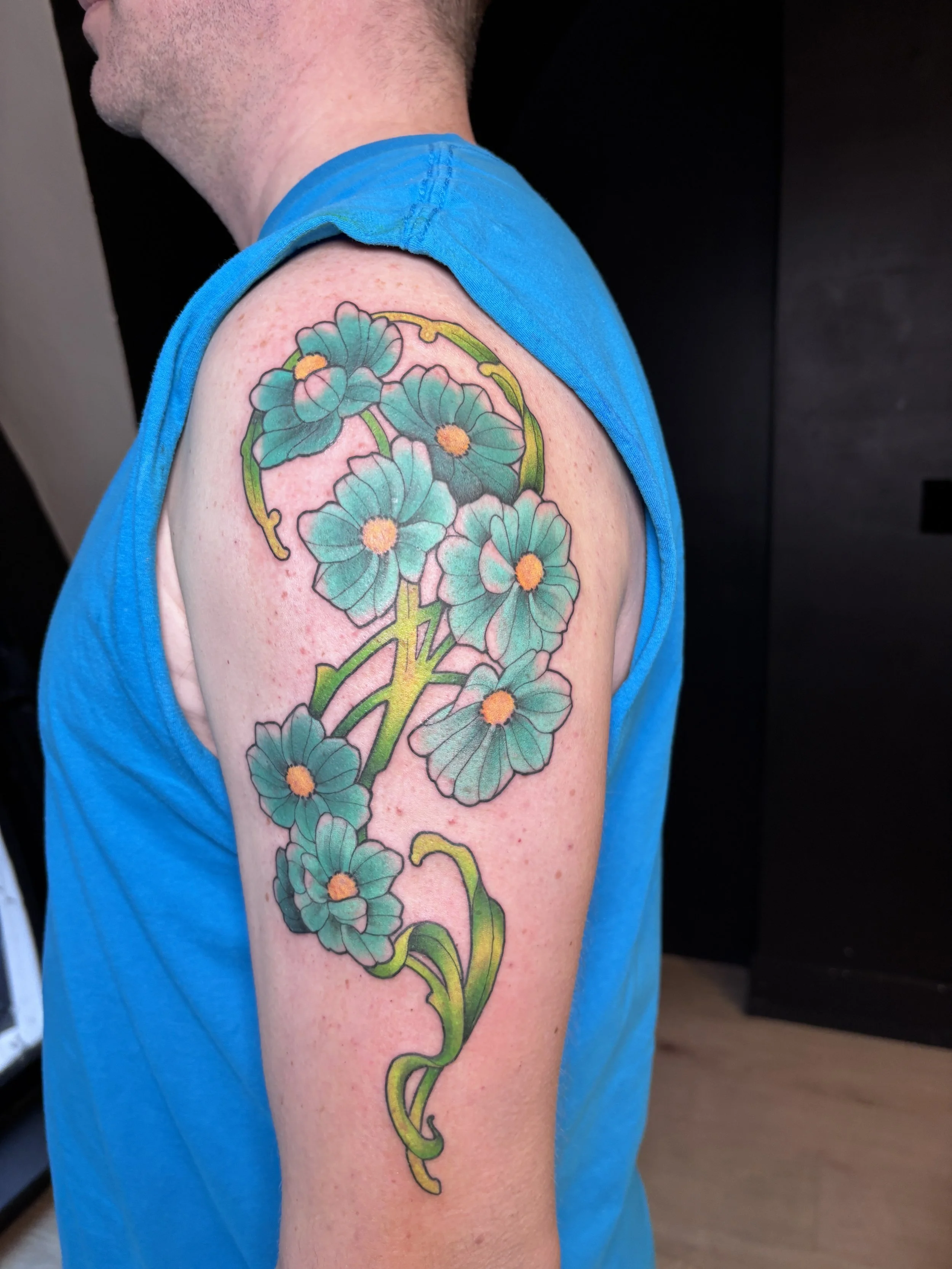 Colorful floral tattoo on a man's upper arm and shoulder, featuring greenish-blue petals with yellow centers and green stems.