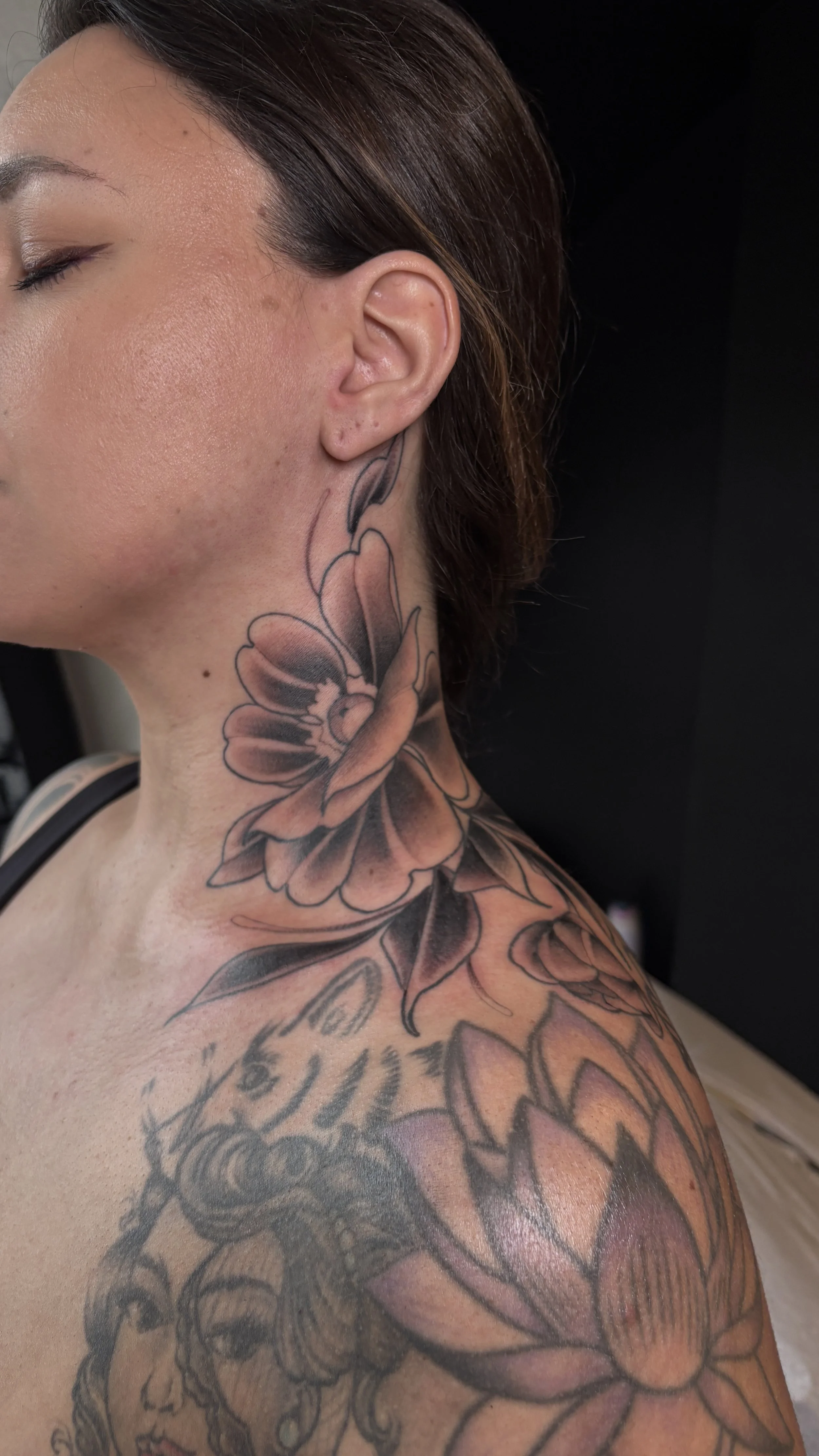 Close-up of a woman's shoulder and neck tattoo featuring large floral designs in black and gray shading.