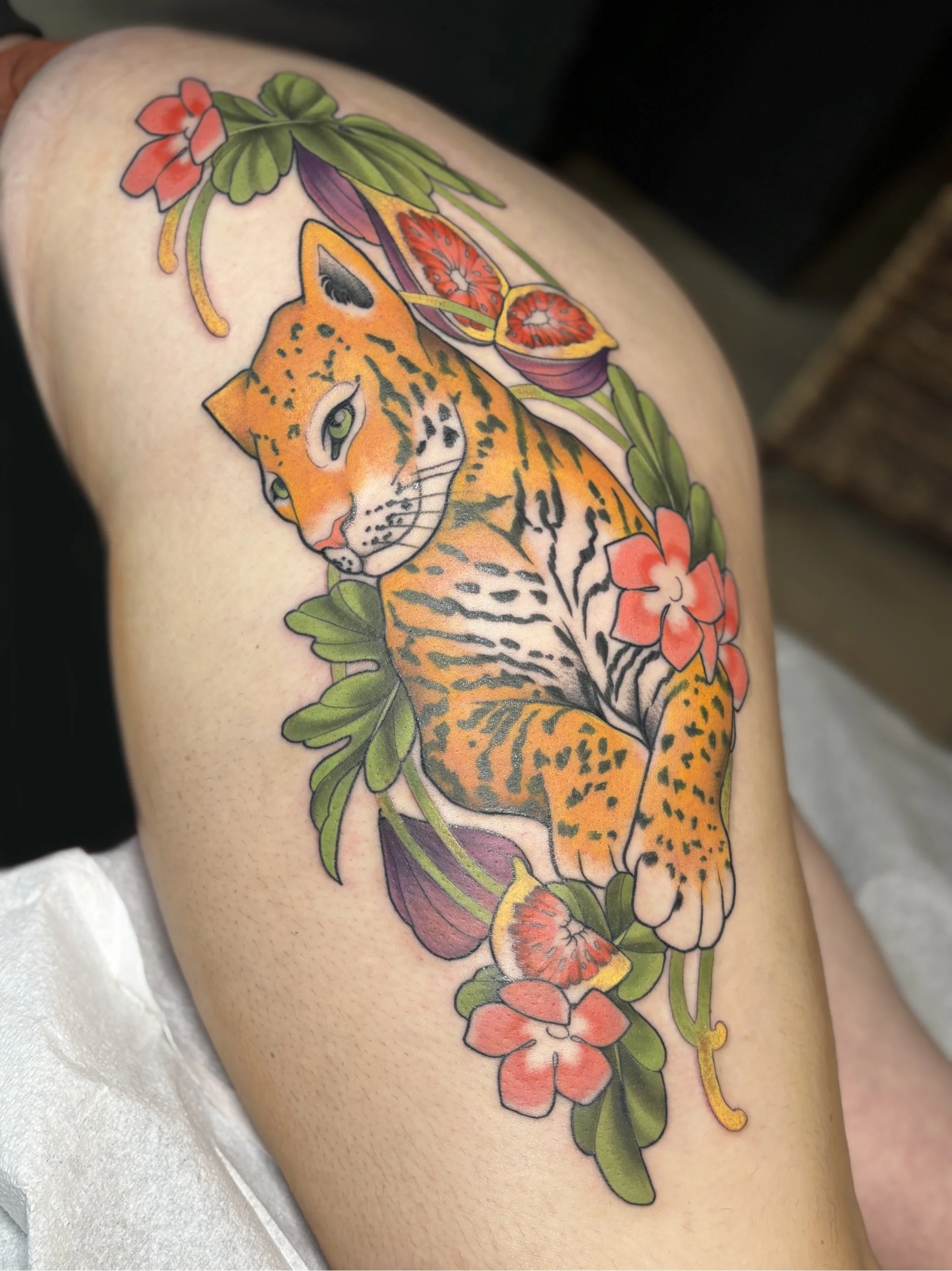 Colorful tattoo of a tiger cub surrounded by pink and purple flowers and green leaves on skin.