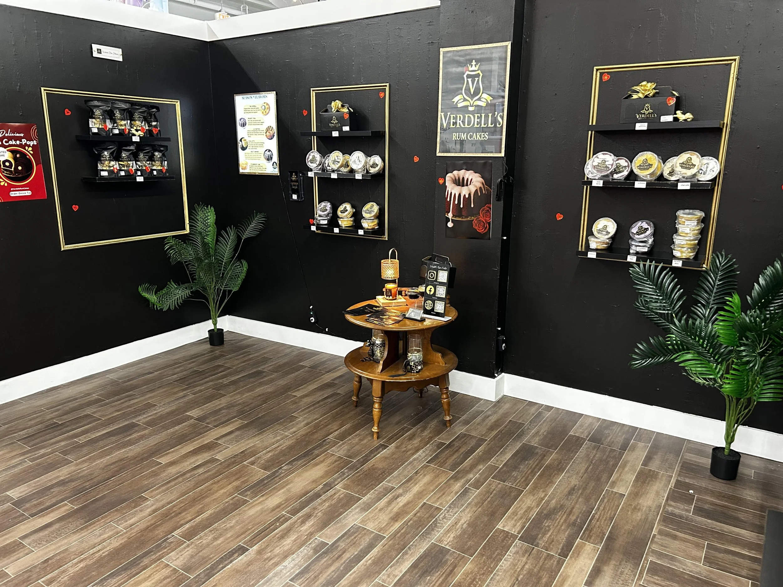 Black wall with decorative shelves displaying various packaged baked goods, two green potted plants, and a small wooden table in front with promotional materials and a lamp, in a store or market setting.