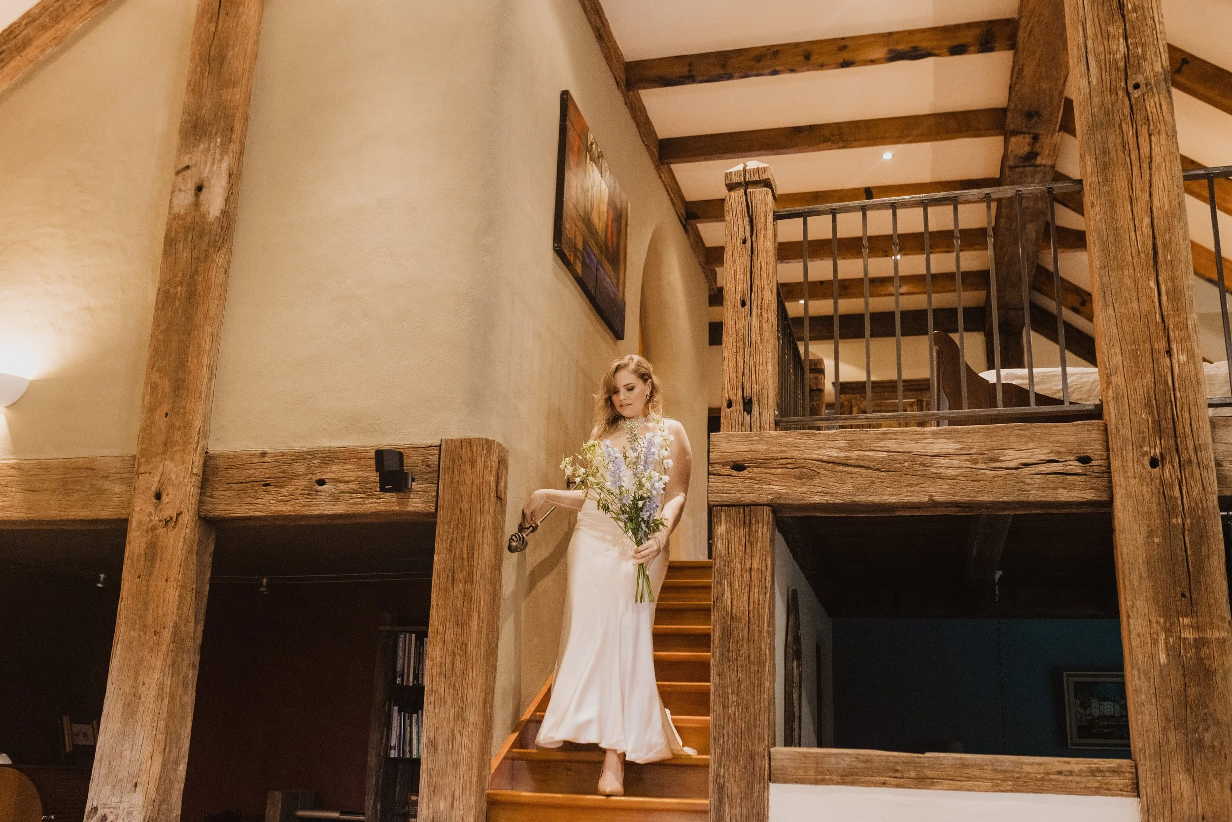 A woman in a white dress holding a bouquet of flowers and a small bell, descending a wooden staircase in a rustic interior with wooden beams and a railing.