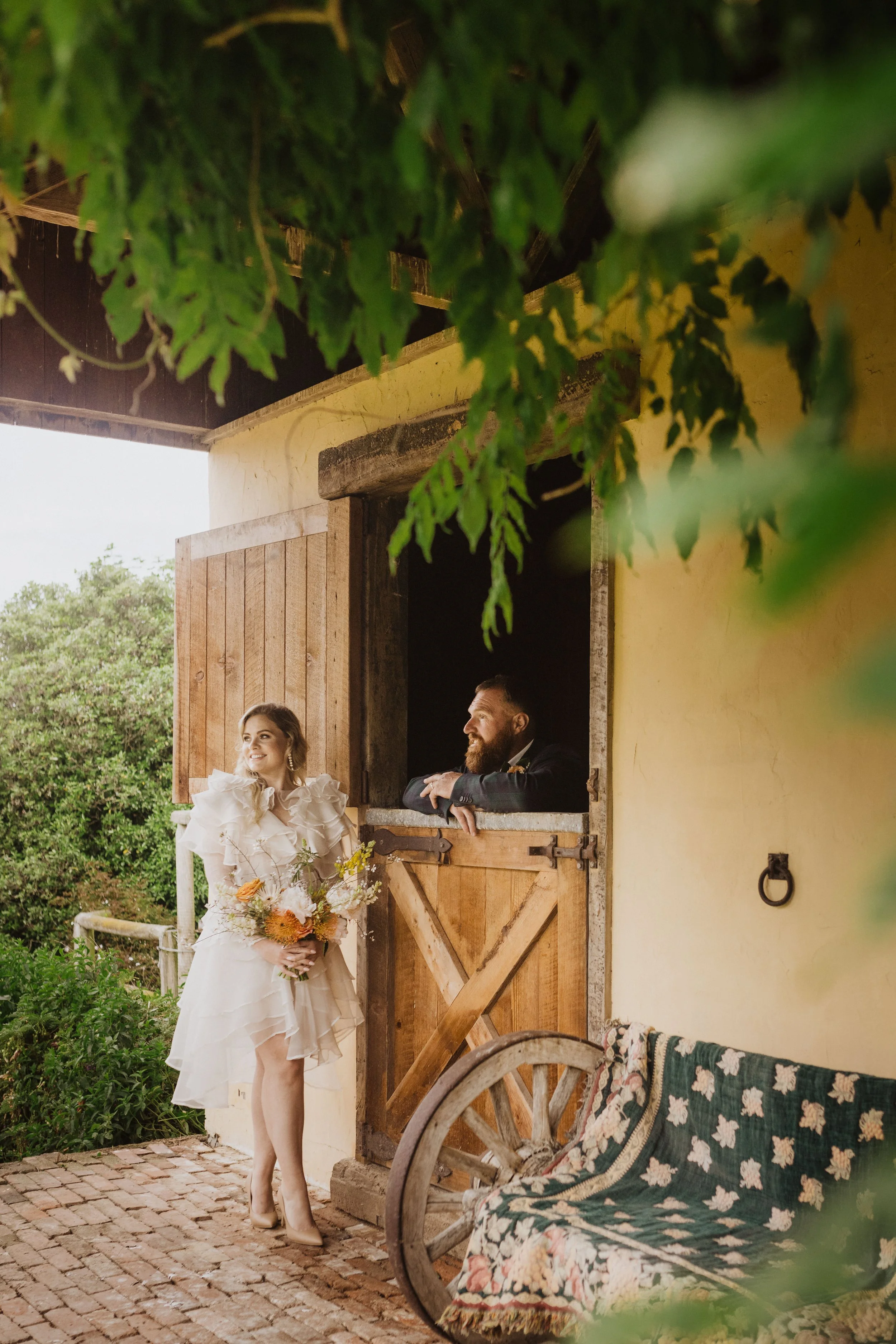 A woman in a white dress holding a bouquet, standing outside a rustic building, looking at a man with a beard leaning out of a small barn window, both smiling.