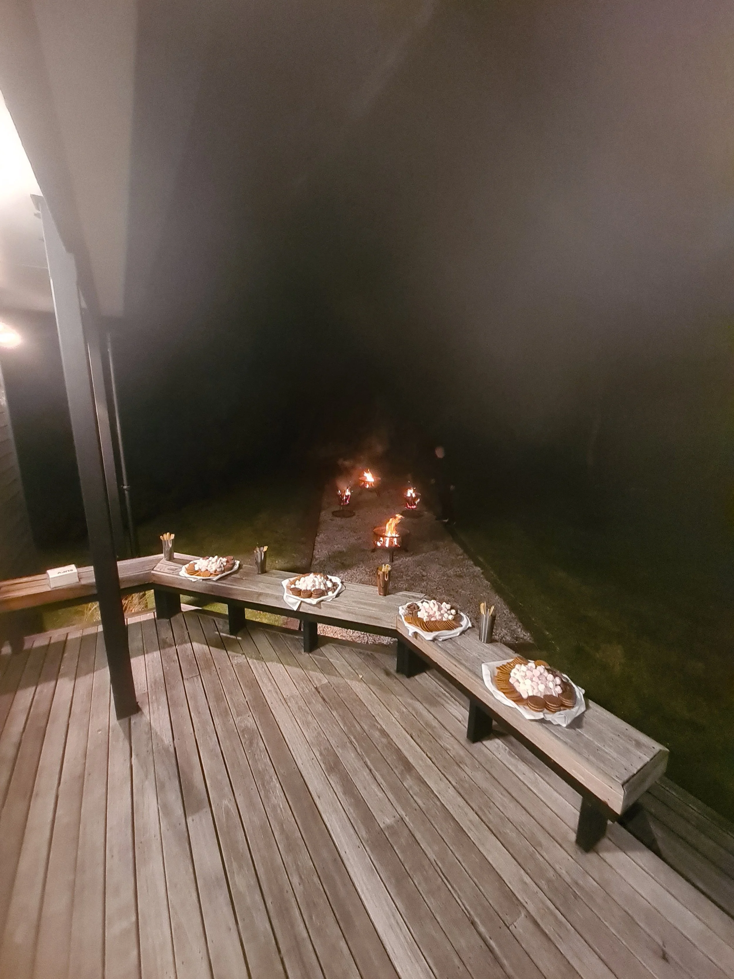 Outdoor celebration setup with plates of cookies and marshmallows on a wooden bench, with small bonfires in the background on a gravel path at night.