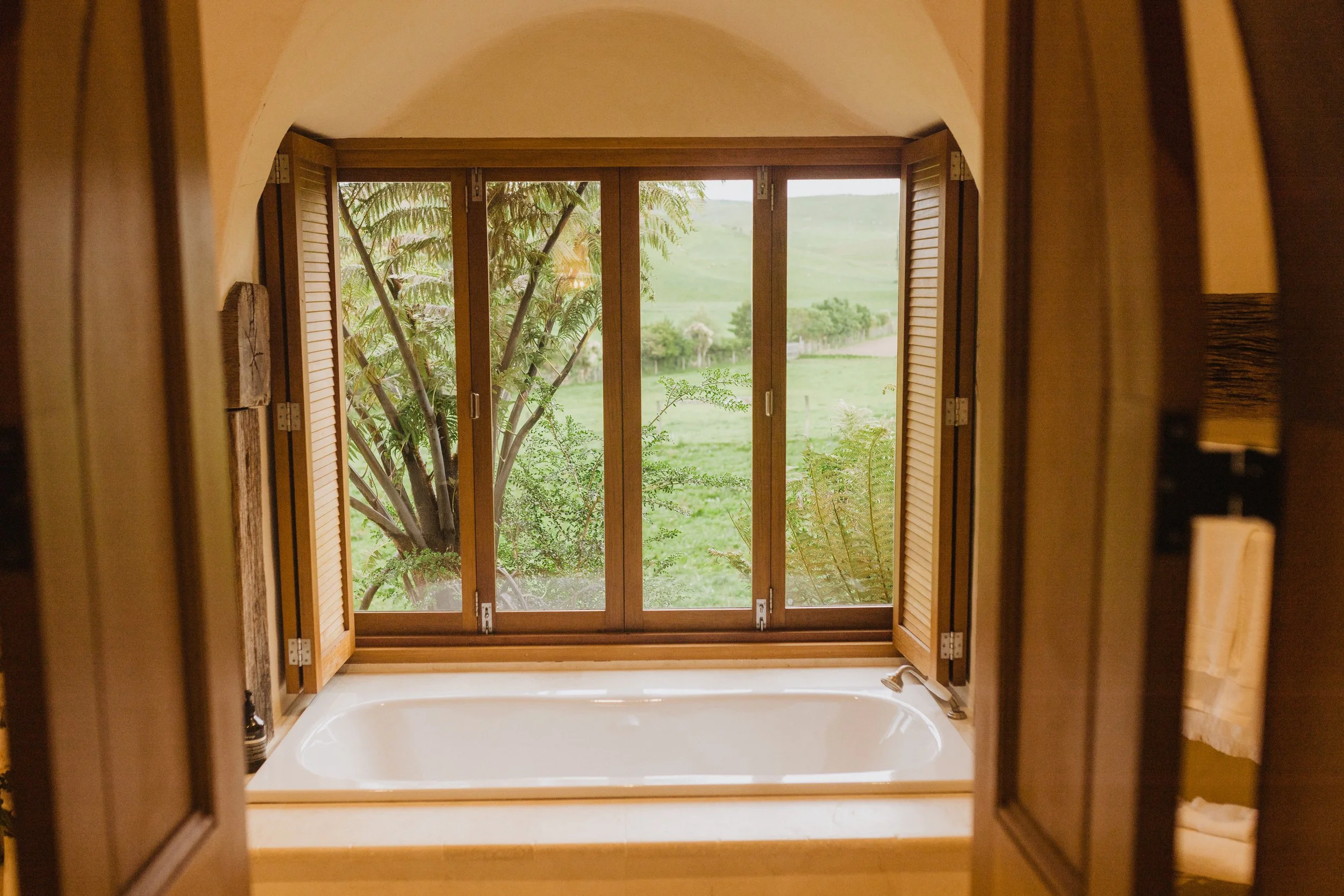 Bathtub with a view of a large green tree through a window with open wooden shutters.