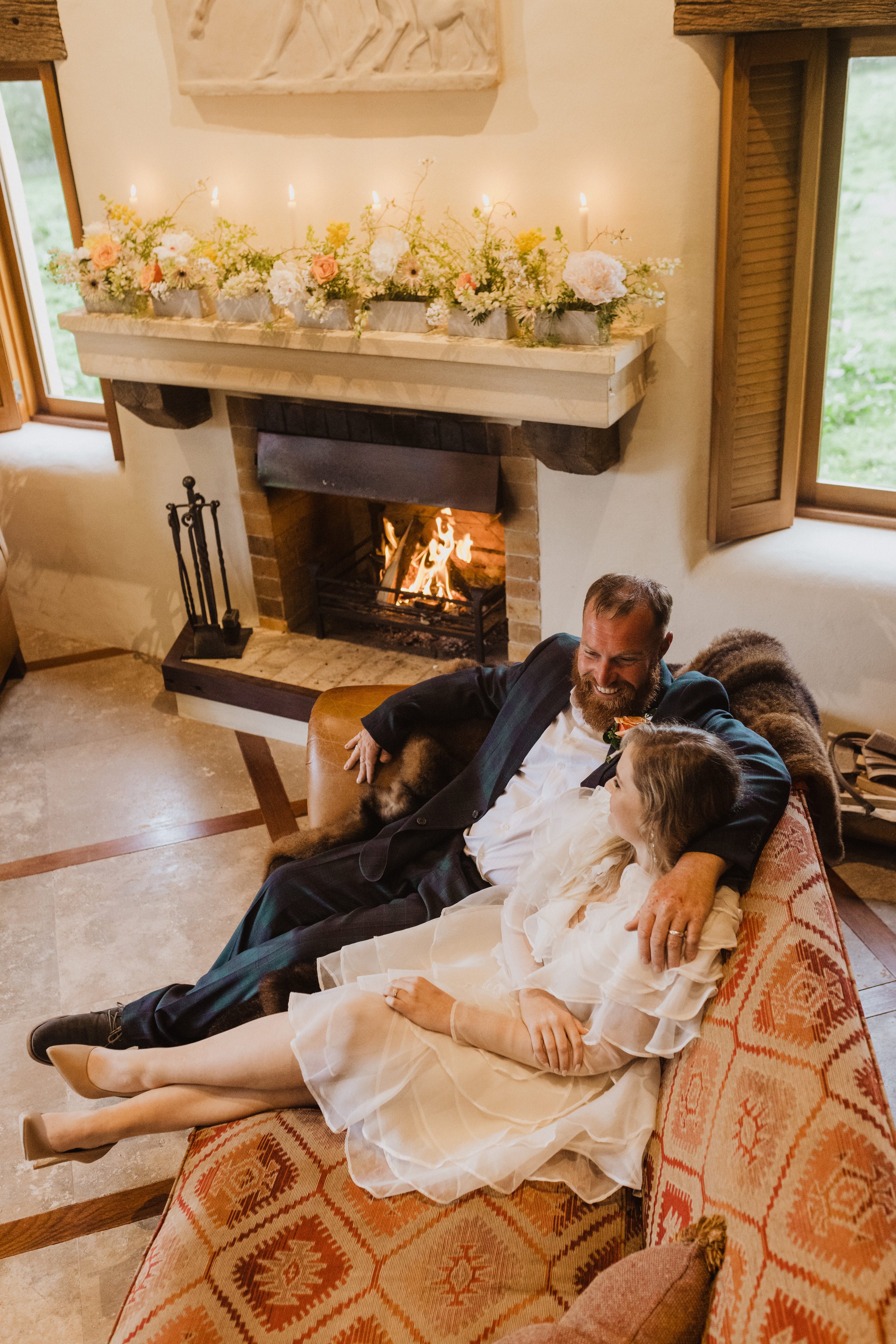 A man and young girl, both dressed in formal attire, sitting together on a patterned sofa in a cozy living room with a fireplace and floral decoration on the mantelpiece.