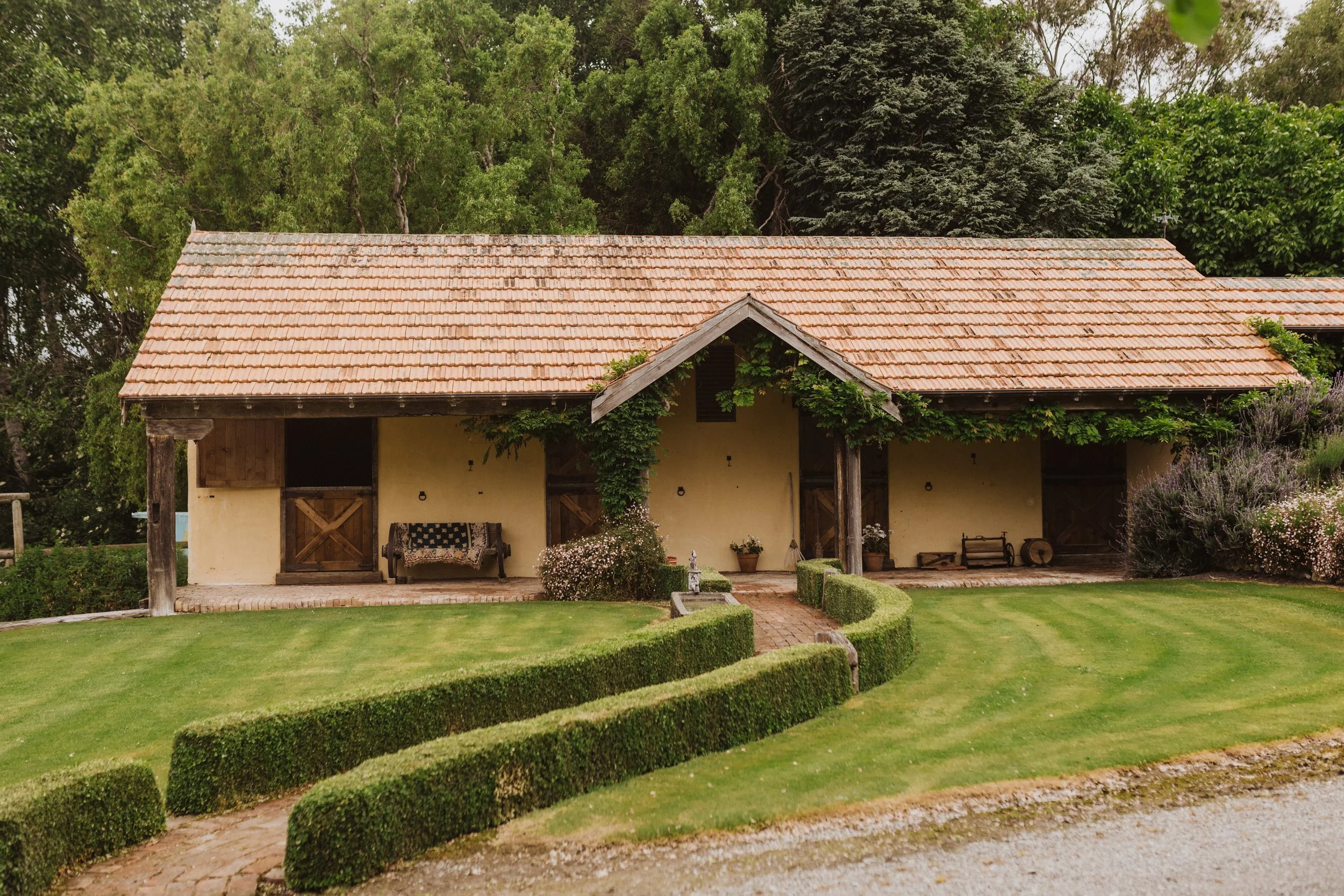 A rustic house with a red-tile roof, surrounded by green trees and a well-kept lawn with trimmed hedges.