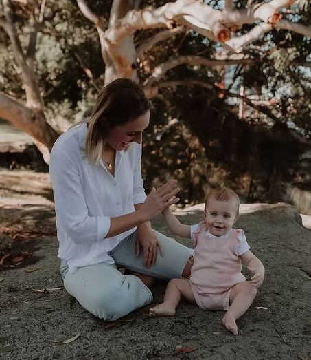 MJ Sleep Consultant, A woman and a toddler girl sitting on a large flat rock outdoors, giving each other a high five, with trees in the background.