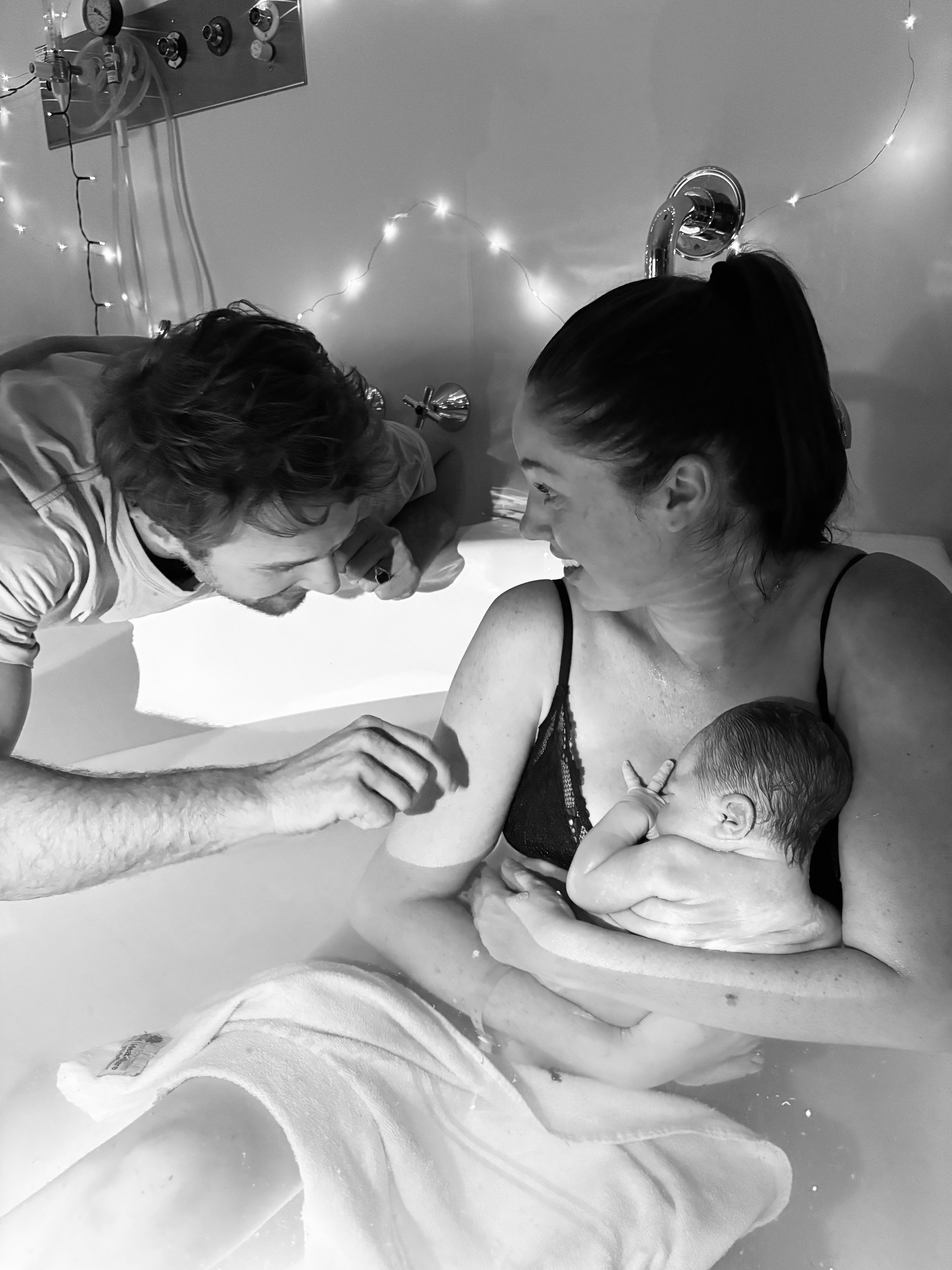 A father reaching out to touch his newborn baby who is in the arms of his mother in a birth pool in hospital surrounded by fairylights.