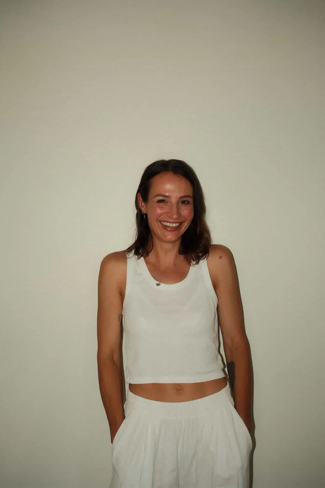 Jade from With Jade Photography, A woman with dark wavy hair, smiling, wearing a white sleeveless top and white pants, standing against a plain light-colored background.