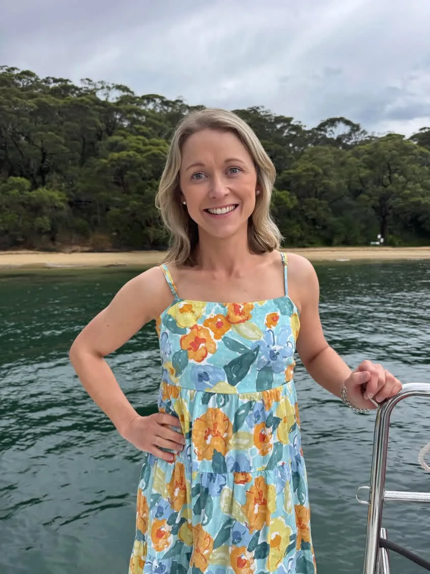Sherree Lopez, a doula from Blossoming Families wearing a colorful floral dress standing on a boat near the water with a forested shoreline in the background, smiling.