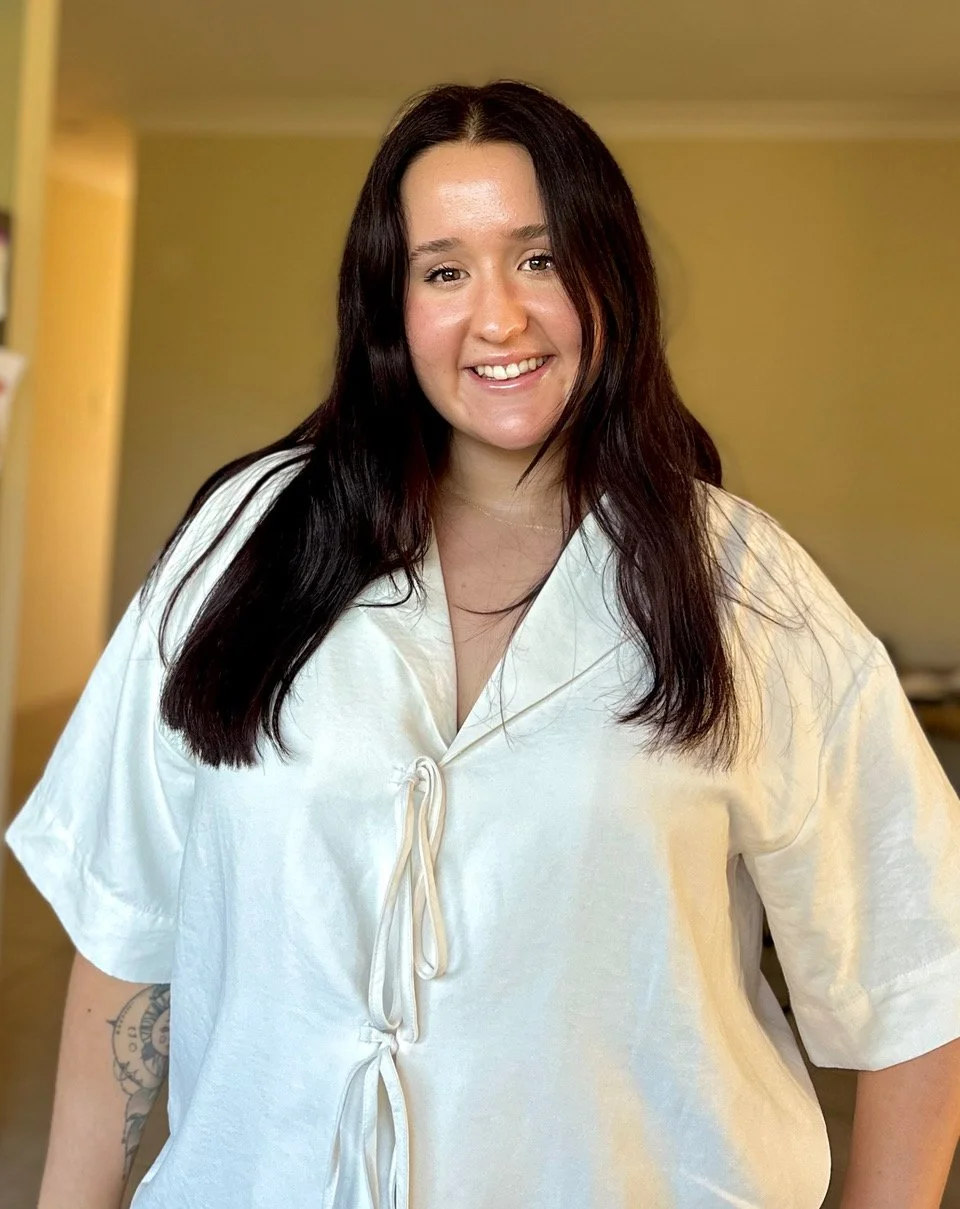 Maegan, from The Gemini Doula, a young woman with long dark hair smiling, wearing a white blouse, standing indoors with a yellow wall background.