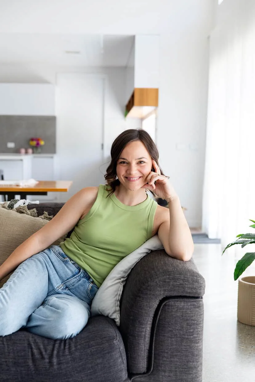 A woman sitting on a dark gray couch in a bright, modern living room, smiling, wearing a light green tank top and light blue jeans, with a white cushion behind her back.