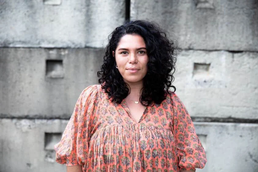 A woman with dark curly hair standing in front of a concrete wall, wearing a floral patterned blouse.