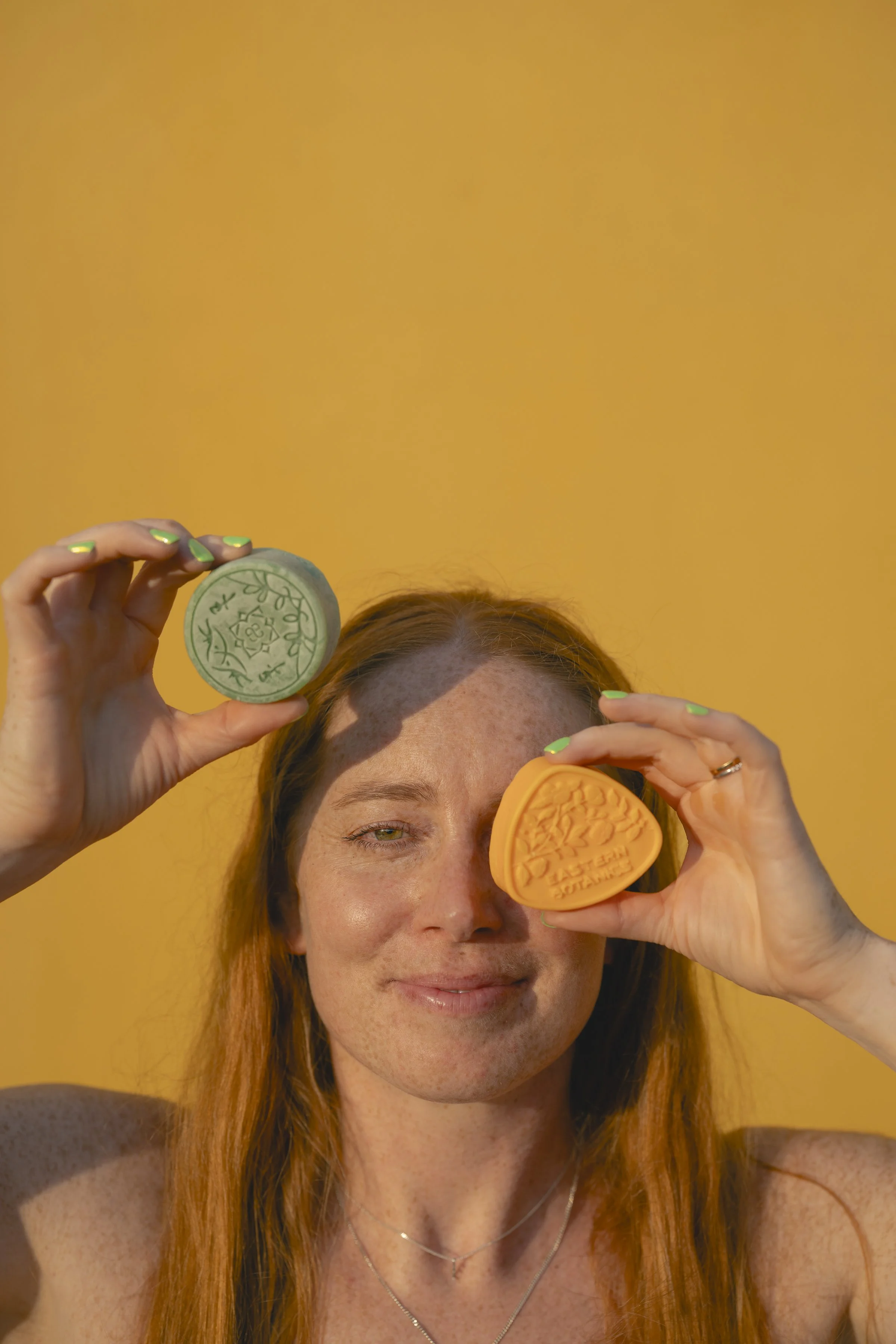 A woman with long red hair holding two colorful round soap bars in front of her face, one green and one orange, with a yellow background.