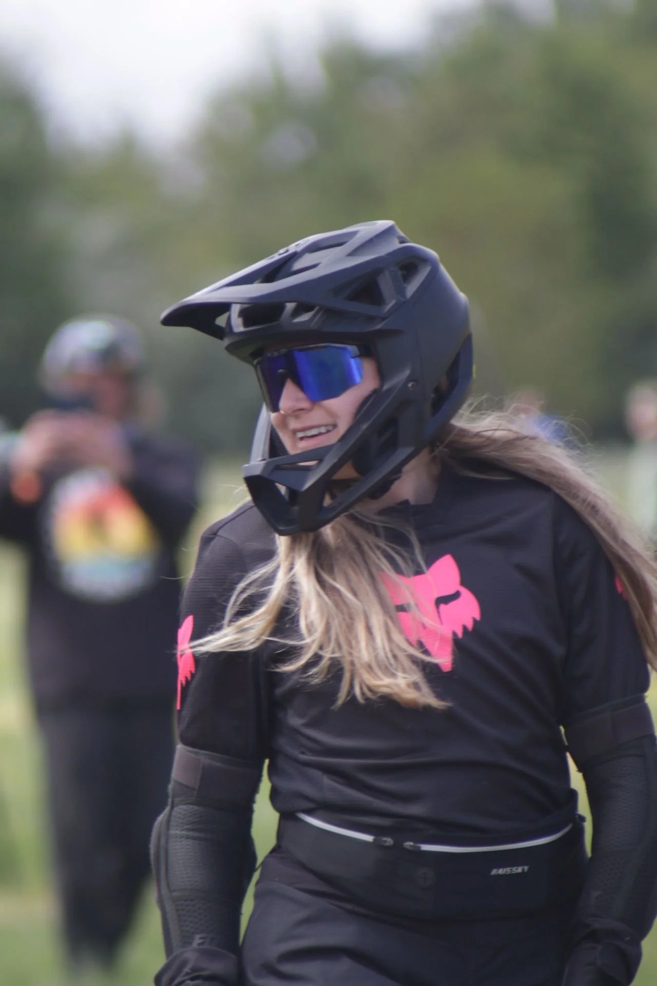 Person wearing a helmet and plaid shirt skateboarding on a dirt trail through a wooded area with green and brown foliage.