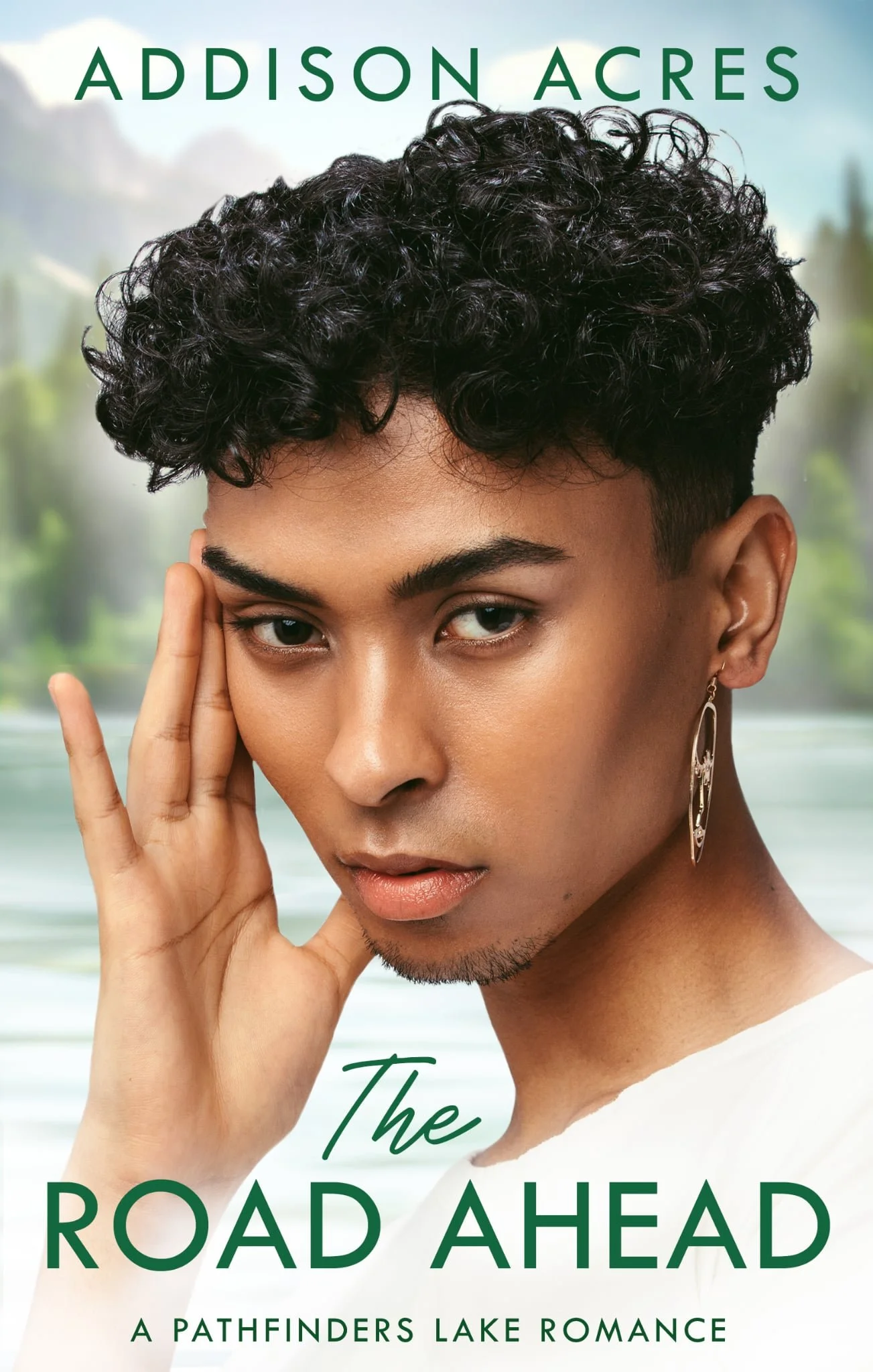 Close-up of a young woman with short, curly black hair, wearing a silver earring, gazing at the camera with her hand gently touching her face, with a blurred nature background.