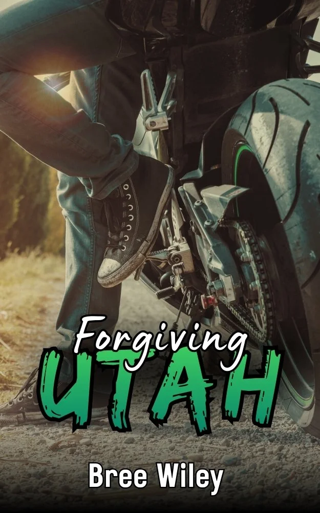 Close-up of a person wearing a black sneaker with white details, standing on a motorcycle footpeg. The motorcycle is parked on a gravel path with greenery in the background. The cover includes the title "Forgiving Utah" and the author's name, Bree Wiley.