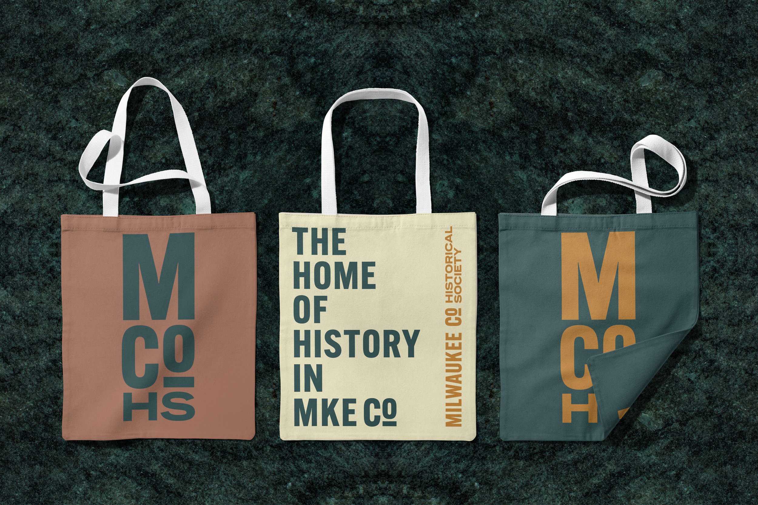Three tote bags with text design, lying on a dark green countertop.