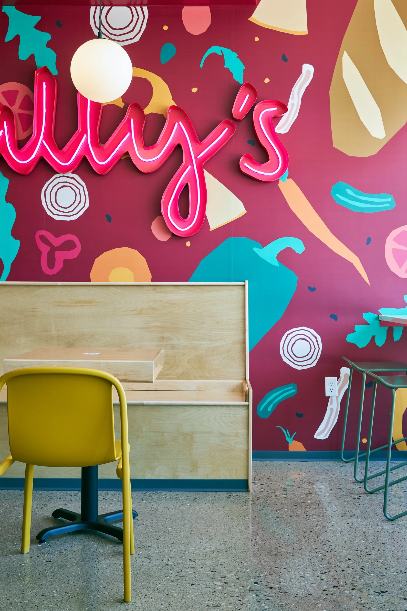 Interior of a colorful cafe or restaurant with a bright pink wall featuring abstract shapes and patterns. There is a yellow chair in the foreground, a light wood counter, and a piece of furniture with green legs to the right.