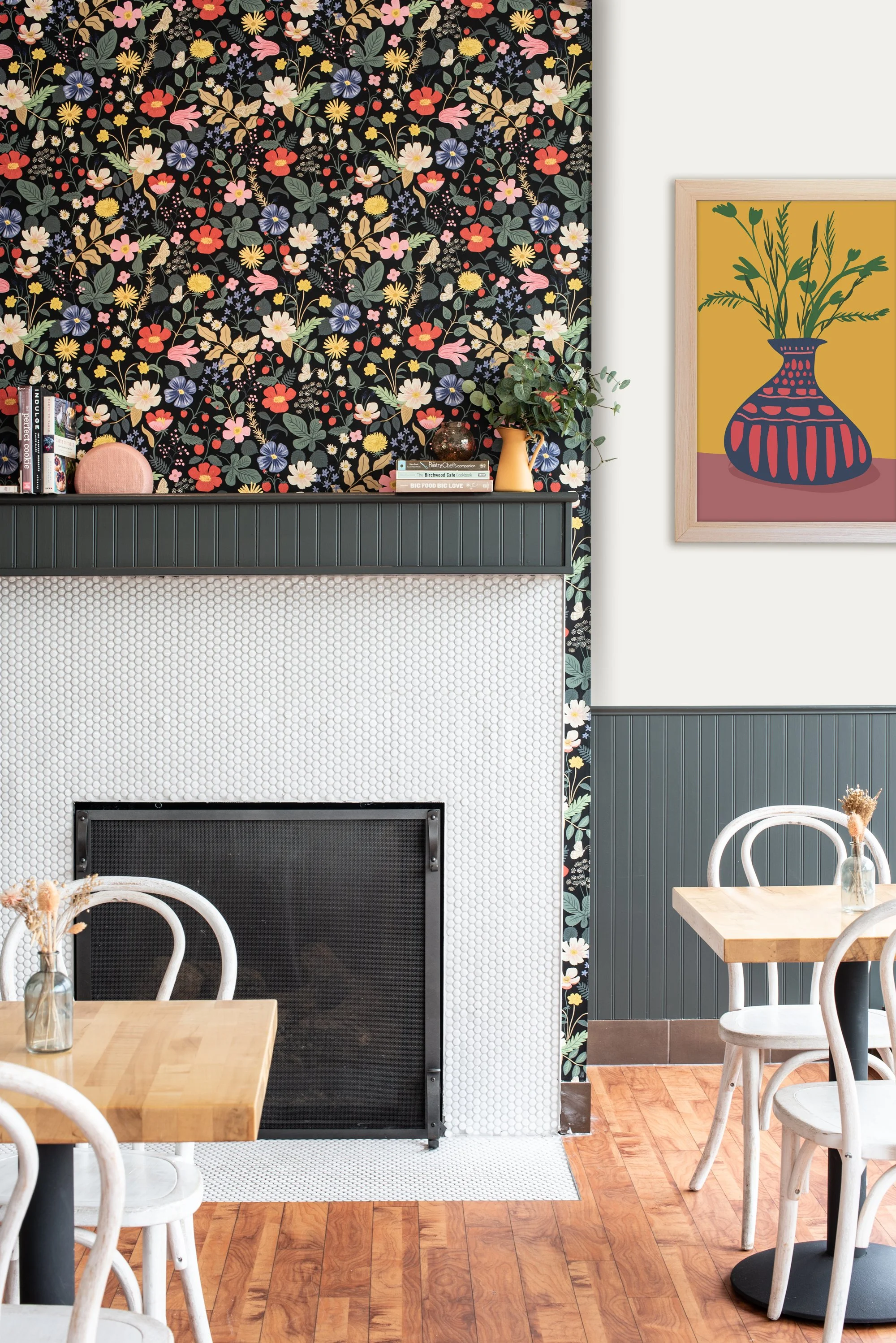 A cozy dining area with wooden tables and white chairs, a black fireplace, a colorful floral wallpaper behind the fireplace, a bar area with books and a plant, and a framed abstract floral painting on the white wall.