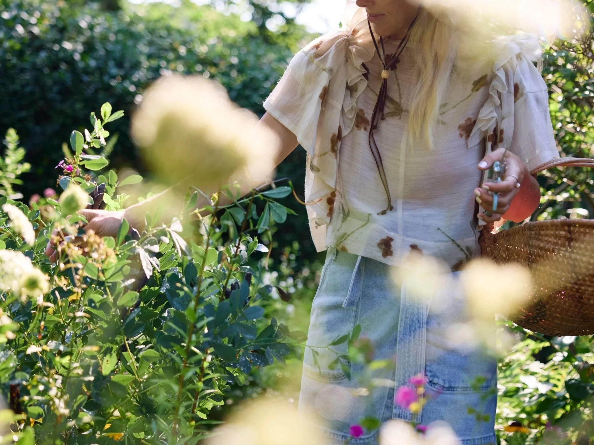 A woman harvesting flowers or plants in a lush garden during daytime, wearing a light-colored blouse and denim shorts, with a basket on her arm.