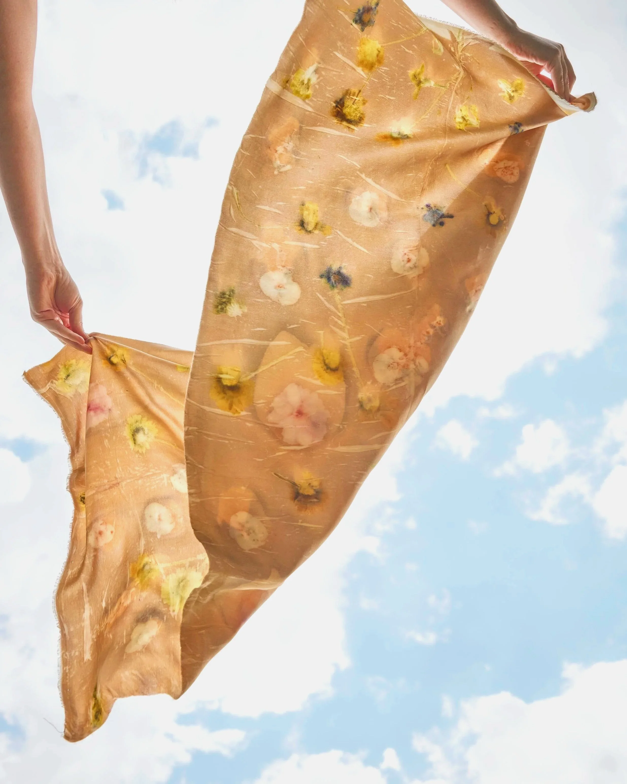 A sheer piece of silk eco-printed with a floral pattern, held up against a partly cloudy sky.